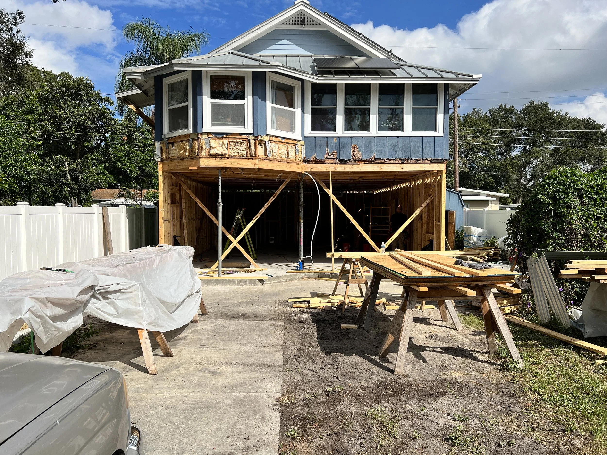A two-story house under construction with the upper level's exterior siding partially installed and the lower level open for future construction. Construction materials and tools are scattered around the site, and a white fence surrounds the backyard.