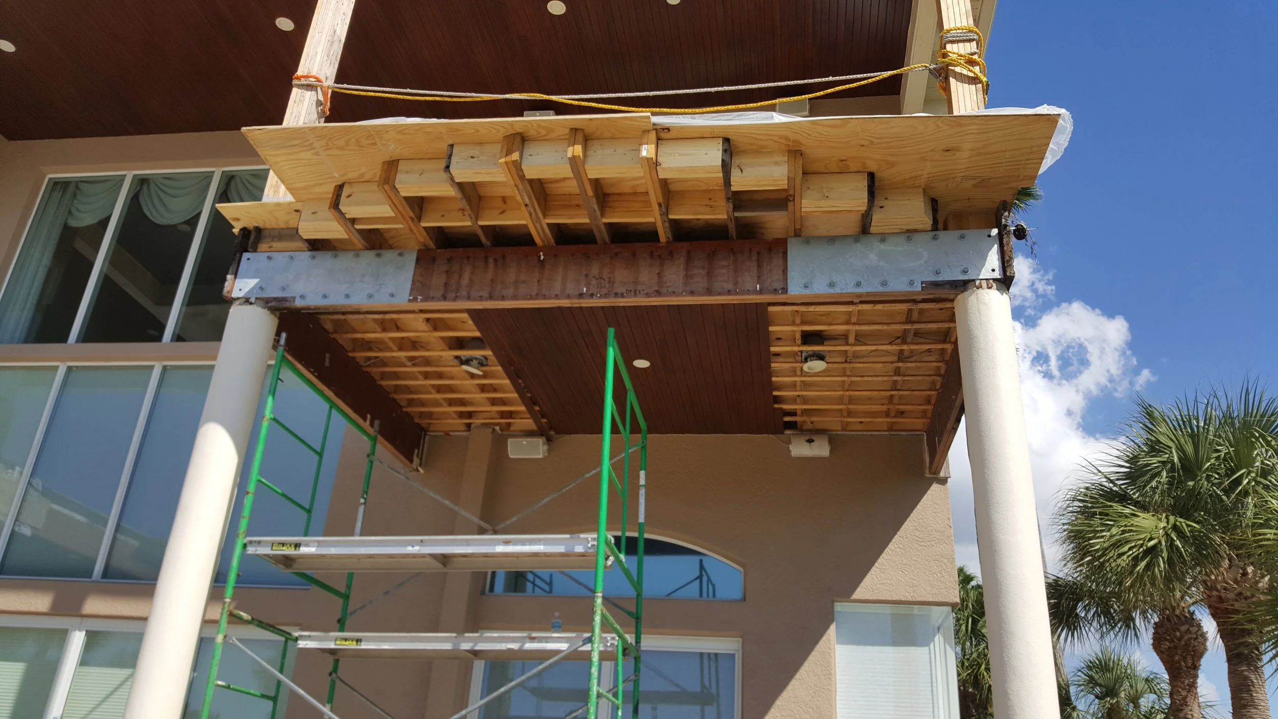 Construction of an outdoor balcony or deck with scaffolding, wooden framing, metal support beam, and a partly finished wooden roof.