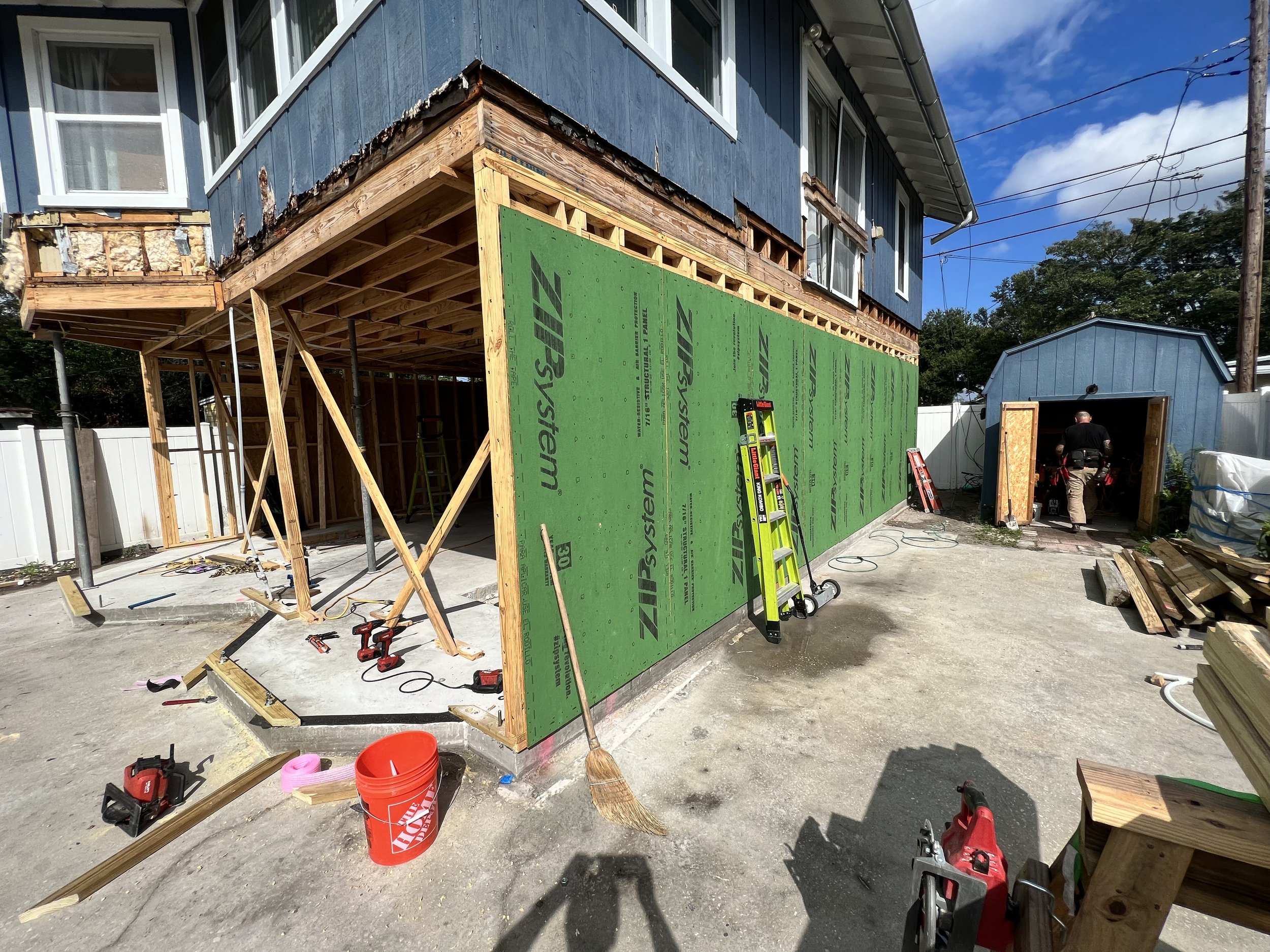 Construction site of a house with siding being installed, showing part of the exterior wall and a garage in the background, tools scattered on the ground.