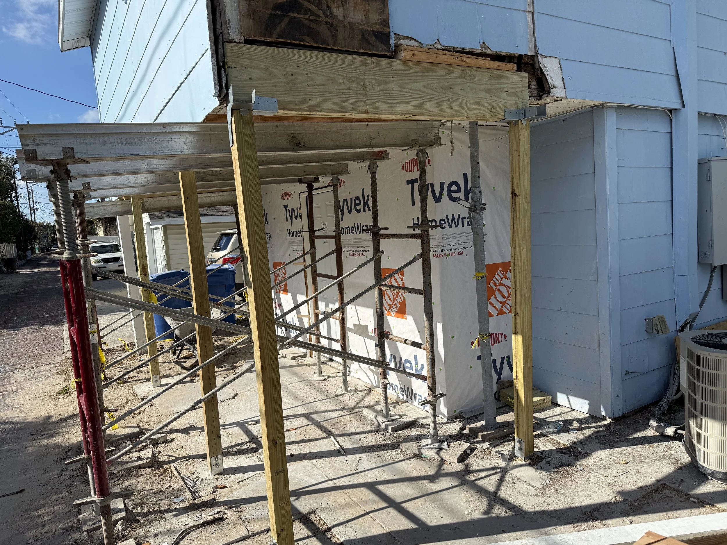 Construction site with scaffolding and exposed building framework attached to a light blue house, dirt ground, neighboring parked cars, and a street with power lines.