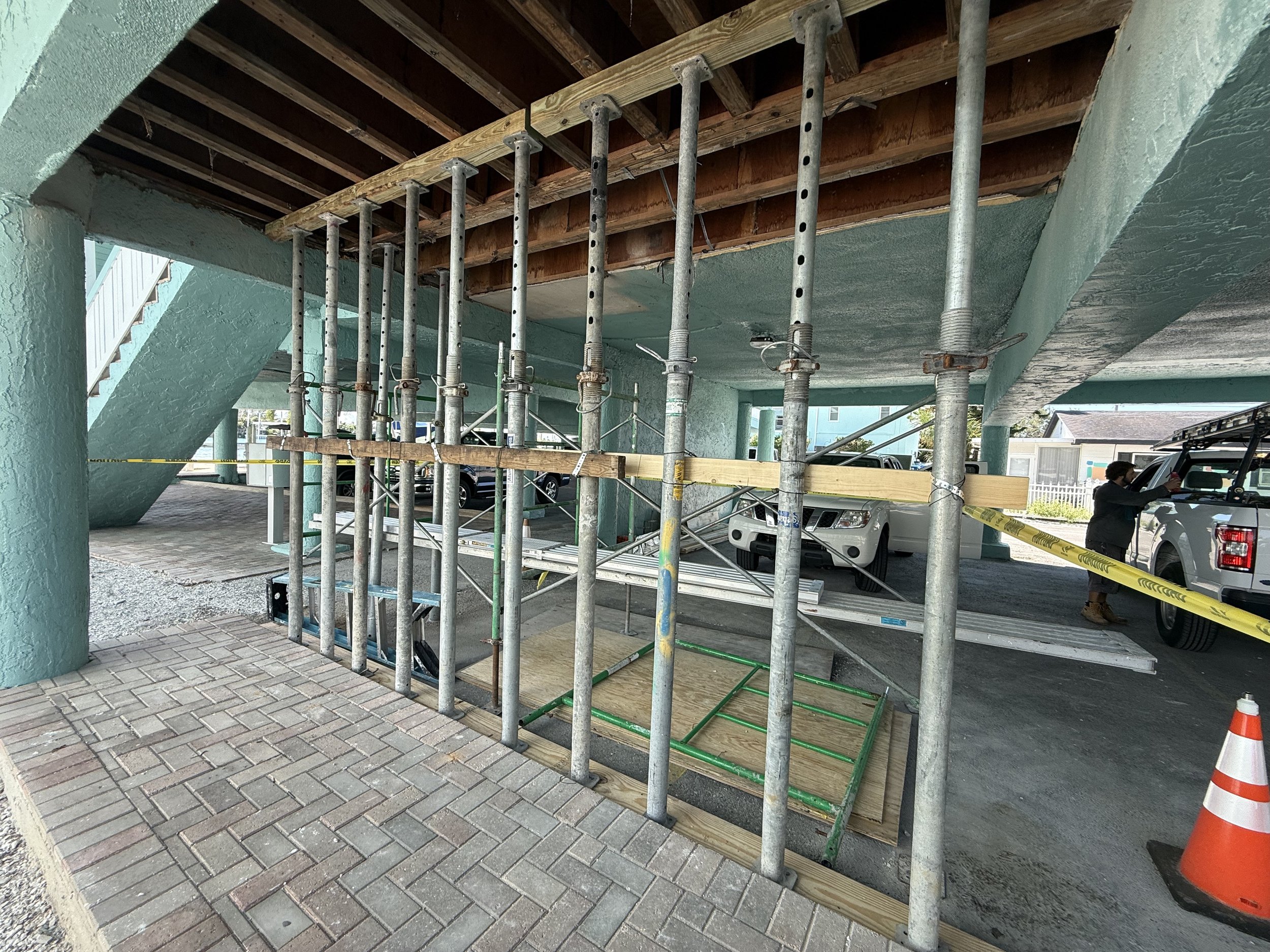 Construction site under a building, with metal scaffolding, wooden planks, and a construction worker near a pickup truck, with orange traffic cone in the foreground.