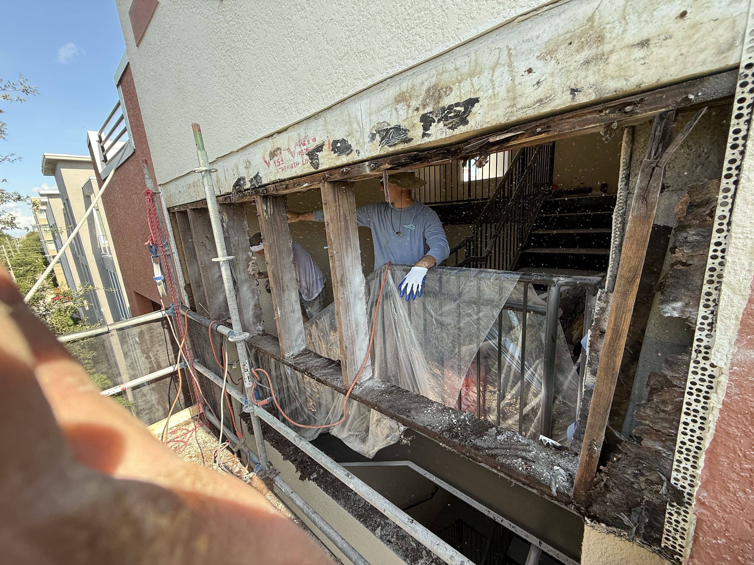 Construction workers renovating the exterior of a building, exposing wooden framing and metal railing, with scaffolding and construction tools visible.