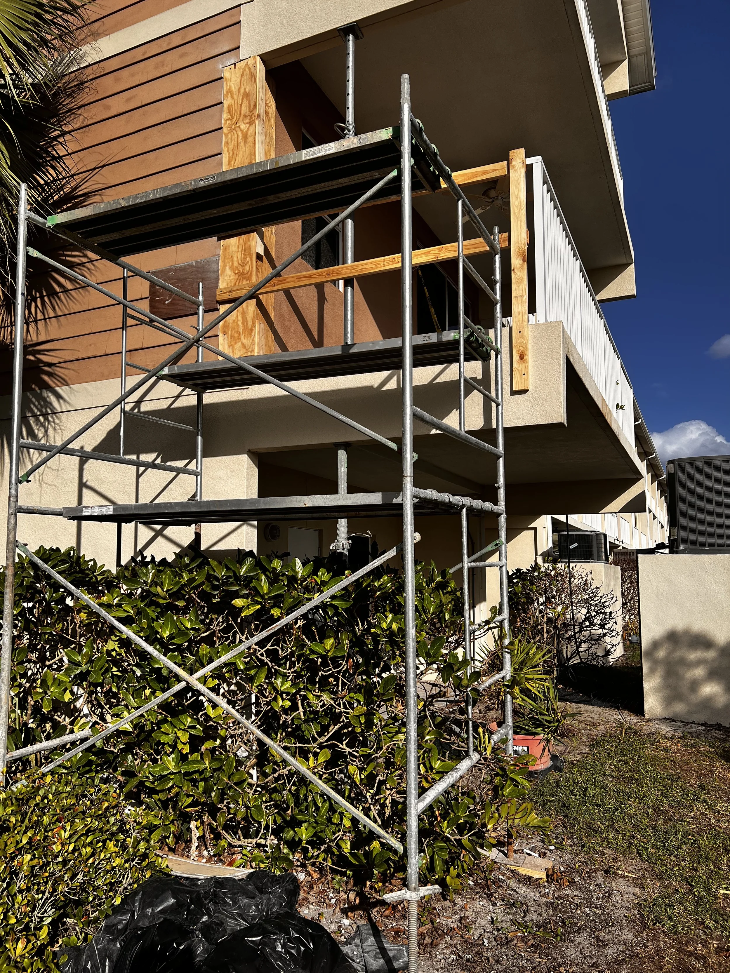 Building exterior under construction with scaffolding in front, beige walls, wooden and white panel siding, and a planter with bushes and a small tree