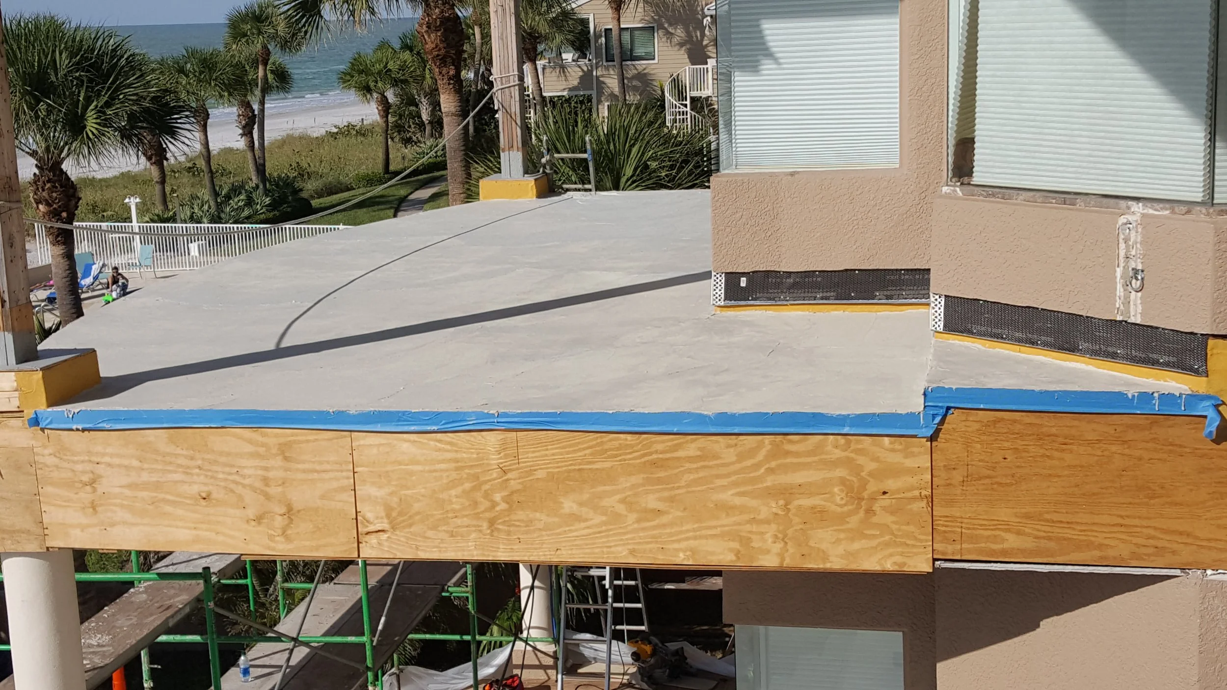 Building under construction with a concrete deck, partially finished beige stucco exterior, and nearby palm trees with a beach and ocean in the background.