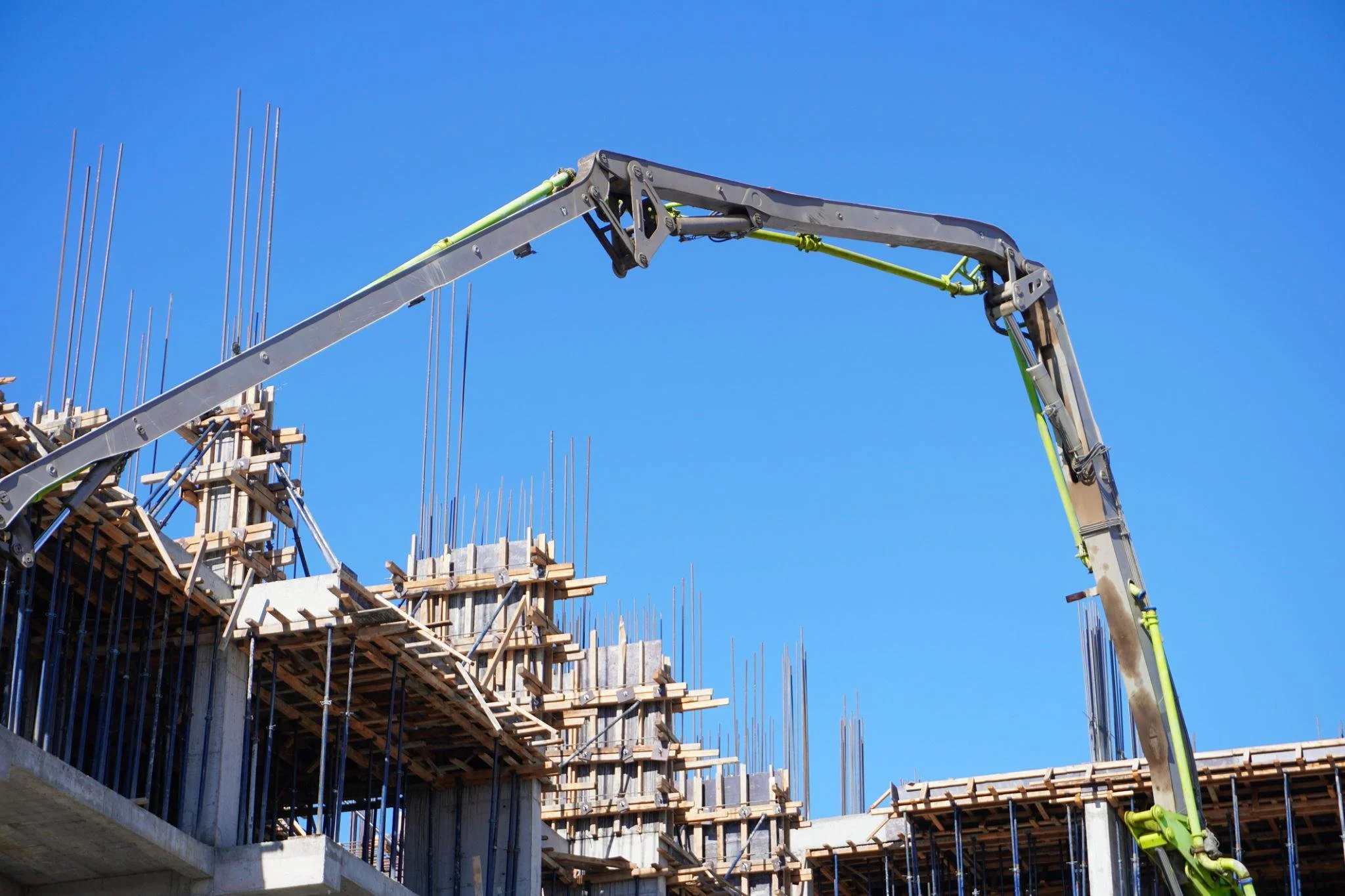 Construction site with concrete structure and wooden formwork, with a large concrete pump placing concrete against a clear blue sky.