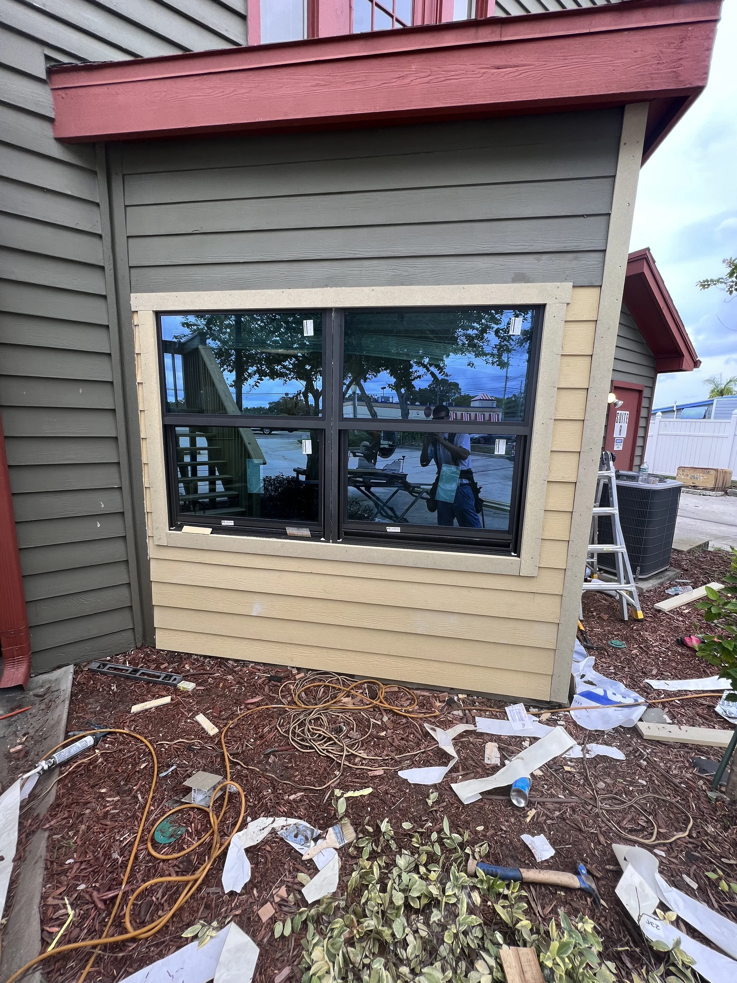 Exterior of a house with beige and green siding, a new black window being installed, construction tools, and debris on the ground.