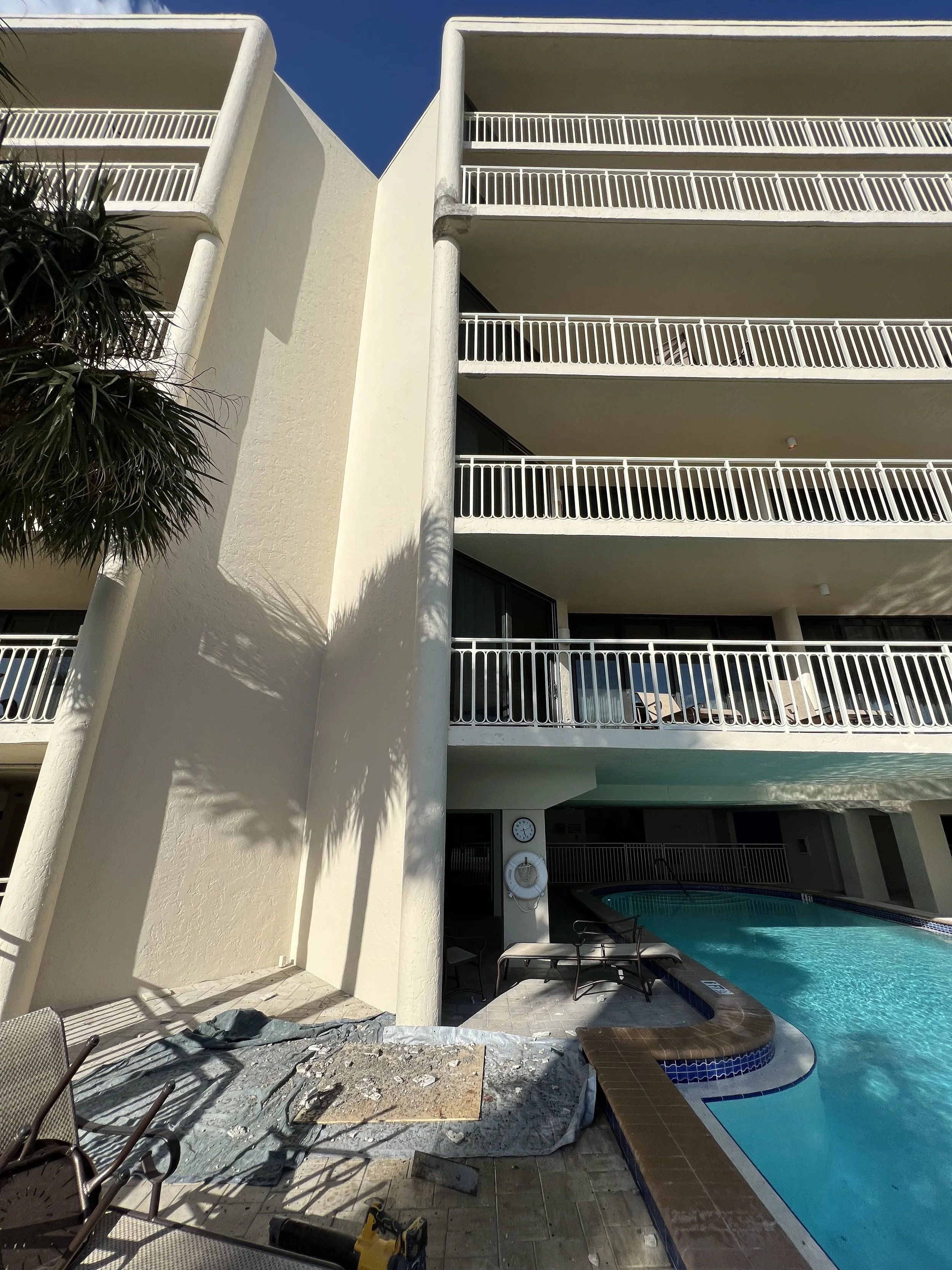 View of a pool area with lounge chairs under a multi-story hotel with white railings and balconies. Part of the construction or repair area with tools and debris is visible in the foreground. Shadows of palm trees are cast on the wall.