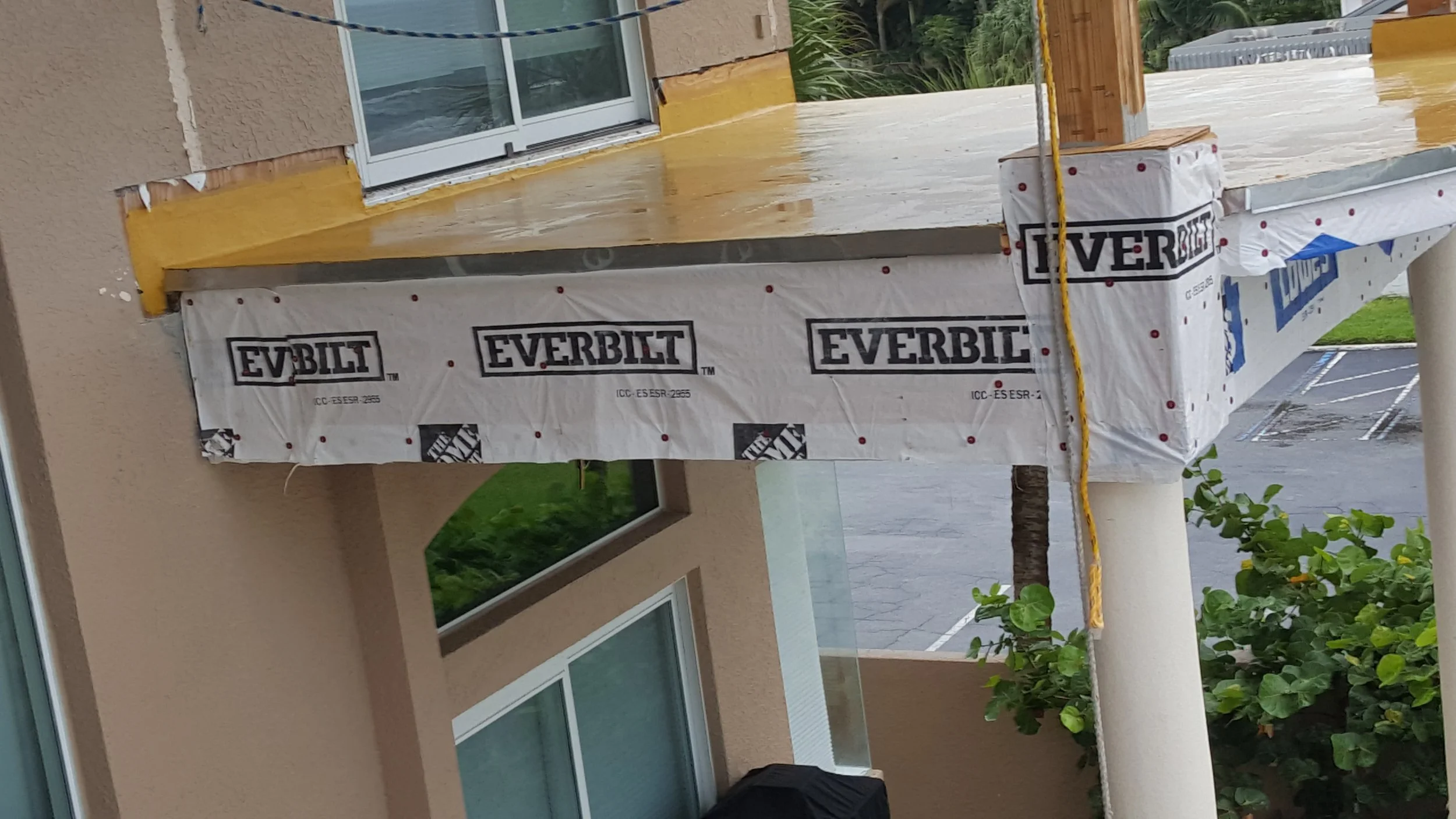 Construction site showing the edge of a building with weatherproofing materials labeled 'Everbuild' and an unfinished deck or balcony area with a sliding glass door, green plants, and parking lot in the background.