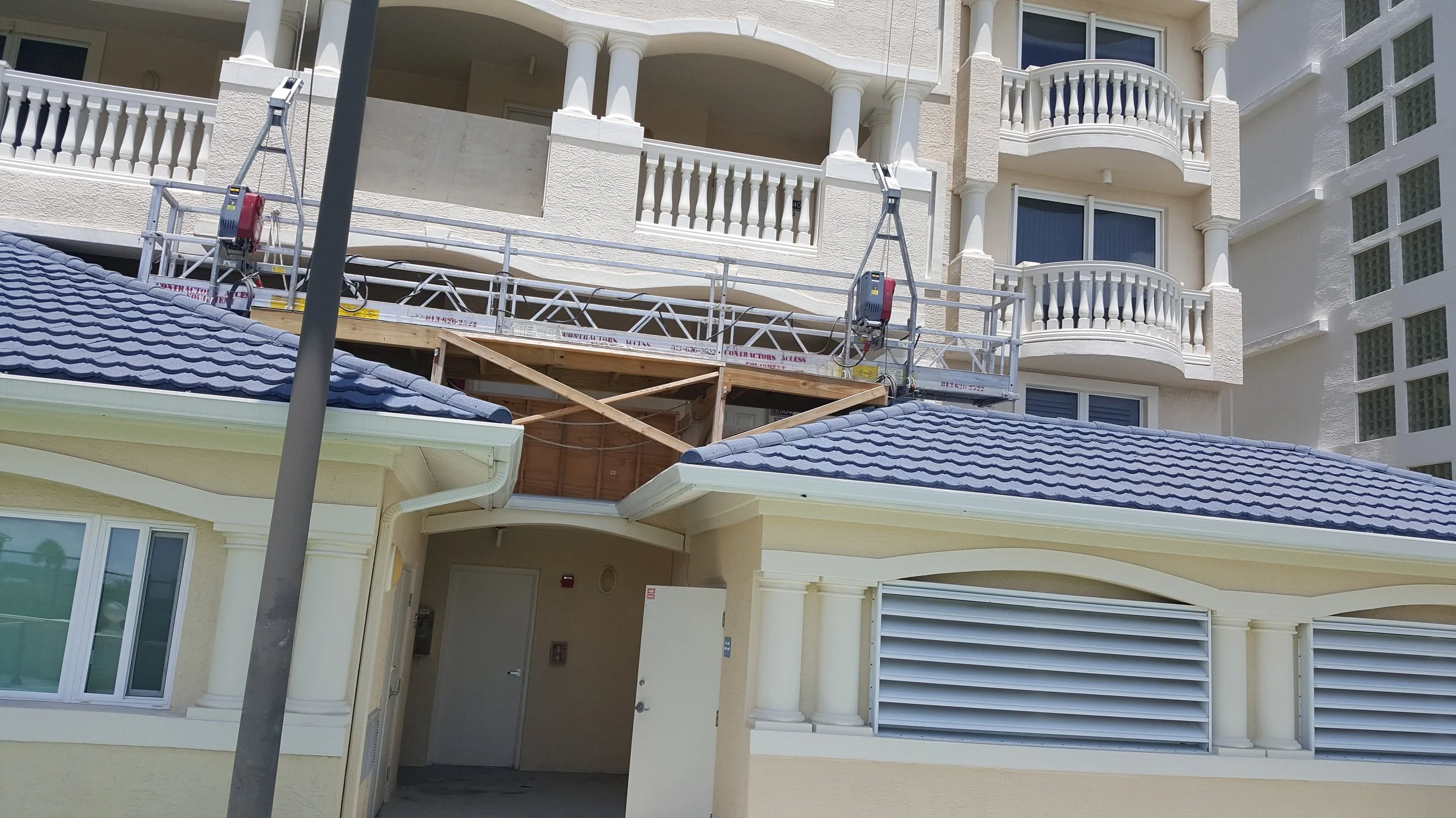 Building under construction with a balcony, scaffolding, and construction equipment on the upper level, and a yellow wall with structural columns and a vent on the lower level.