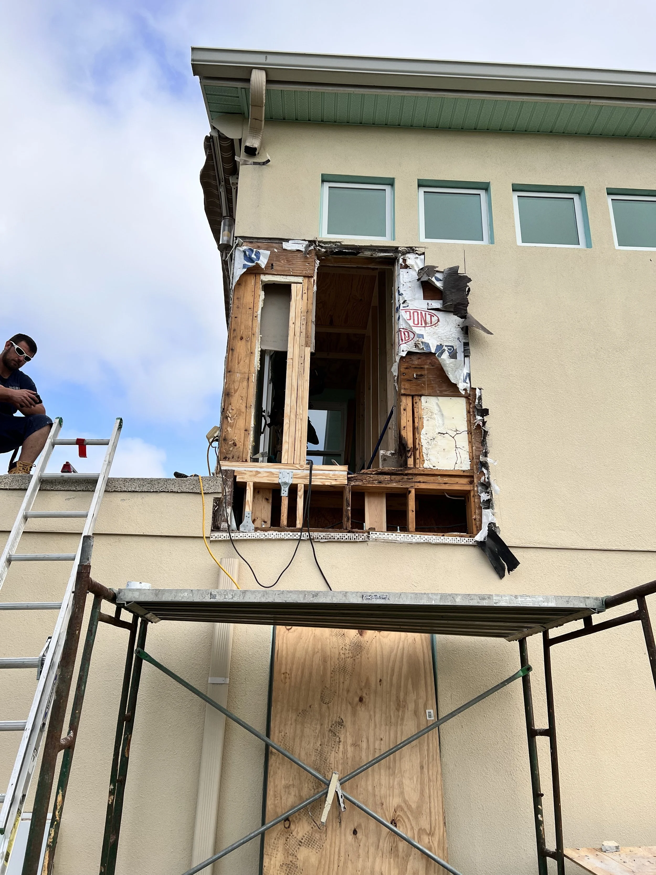 House under renovation with an open wall exposing wooden framing. A worker sits on a ladder to the left, and scaffolding is below the opening.