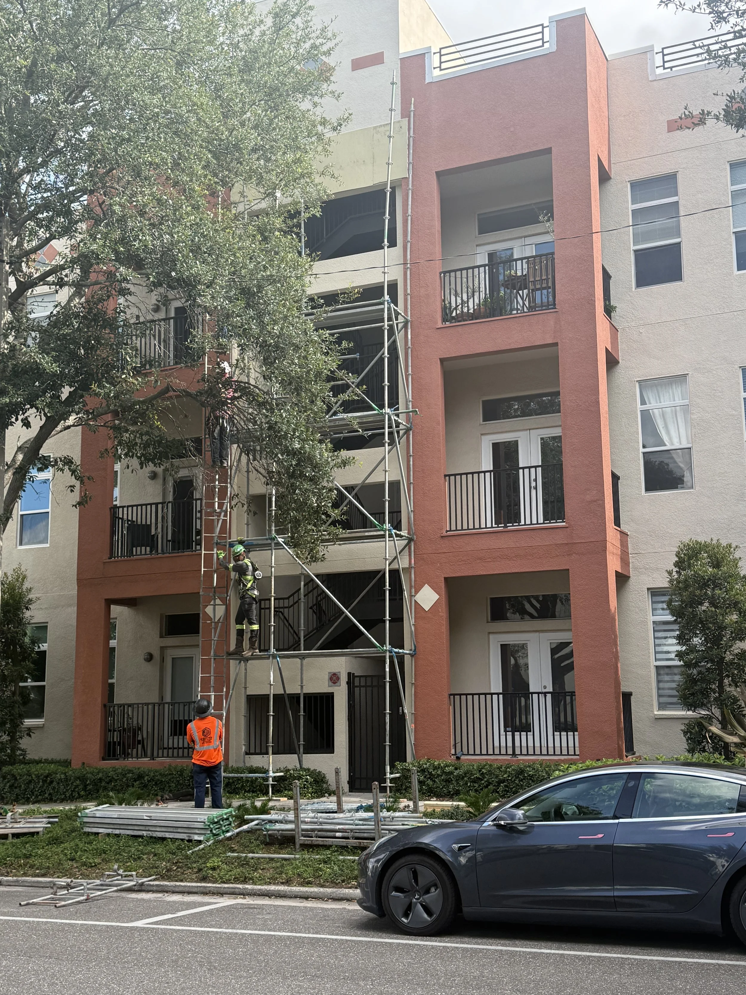 Construction workers assembling scaffolding outside a multi-story residential building with balconies, with a parked car in the foreground.