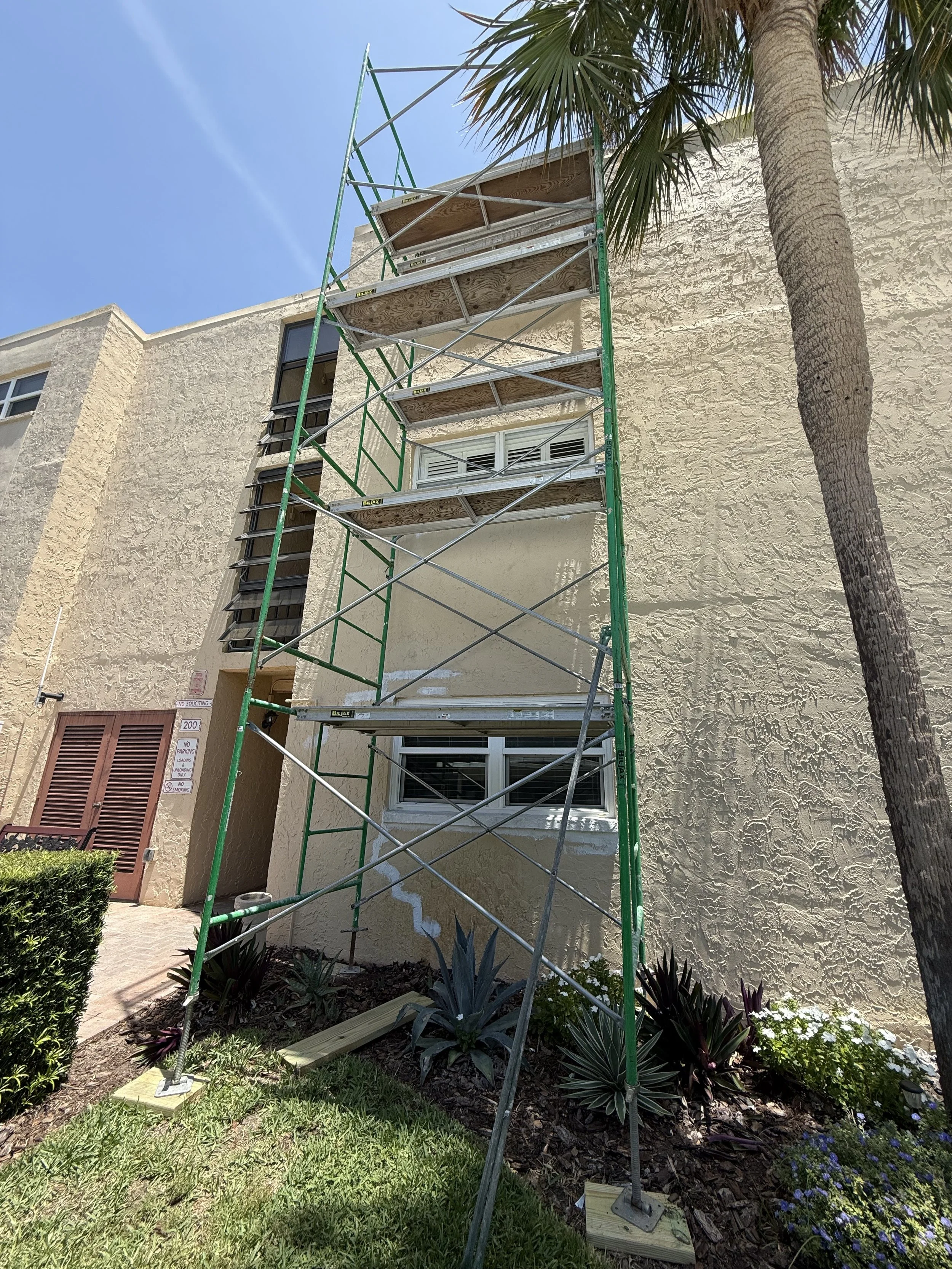 Construction scaffolding set up beside a beige stucco building with windows, surrounded by landscaping including palm trees and plants.