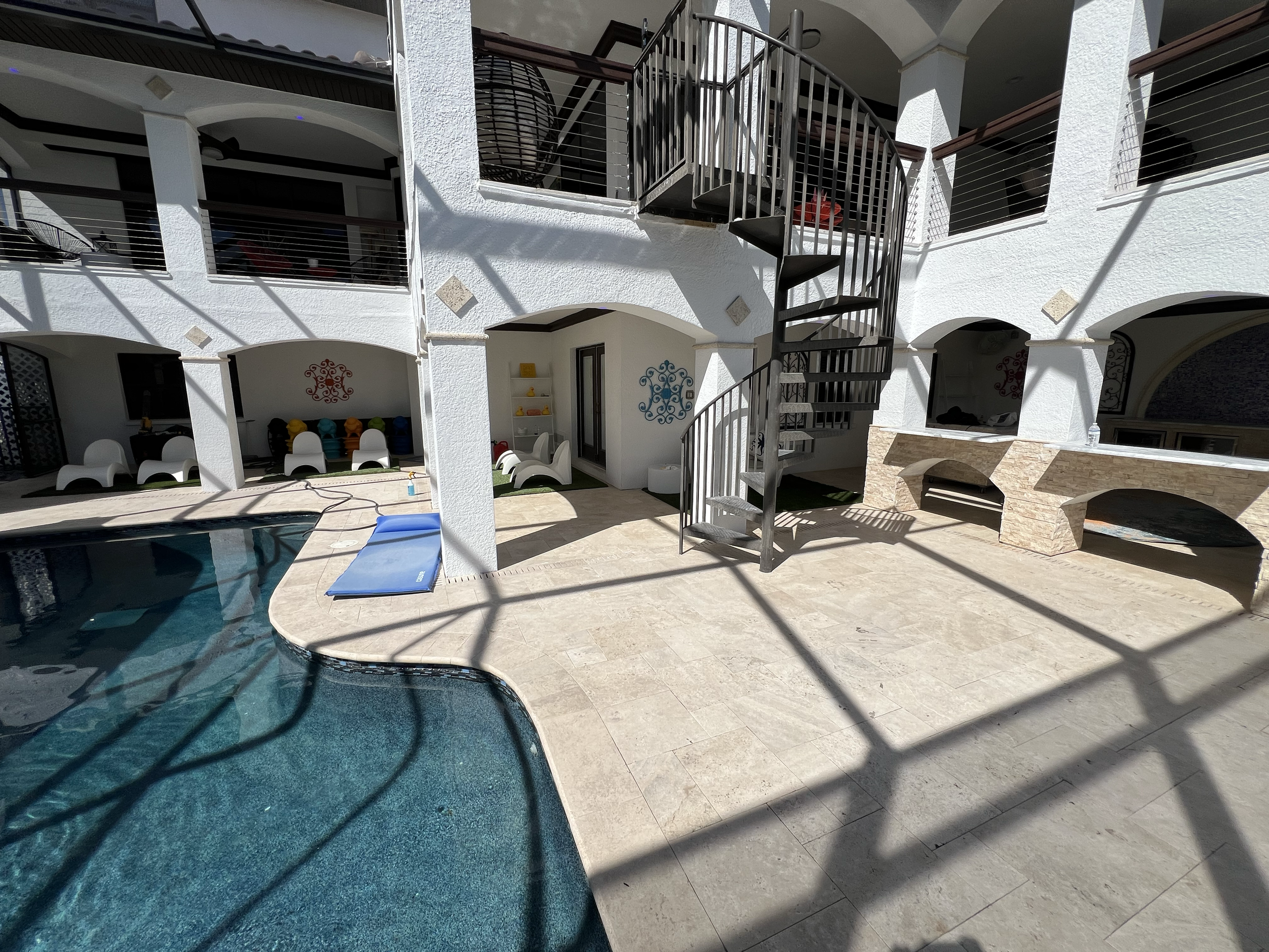 Poolside patio area with spiral staircase, lounge chairs, and arched openings in a white building with balconies.