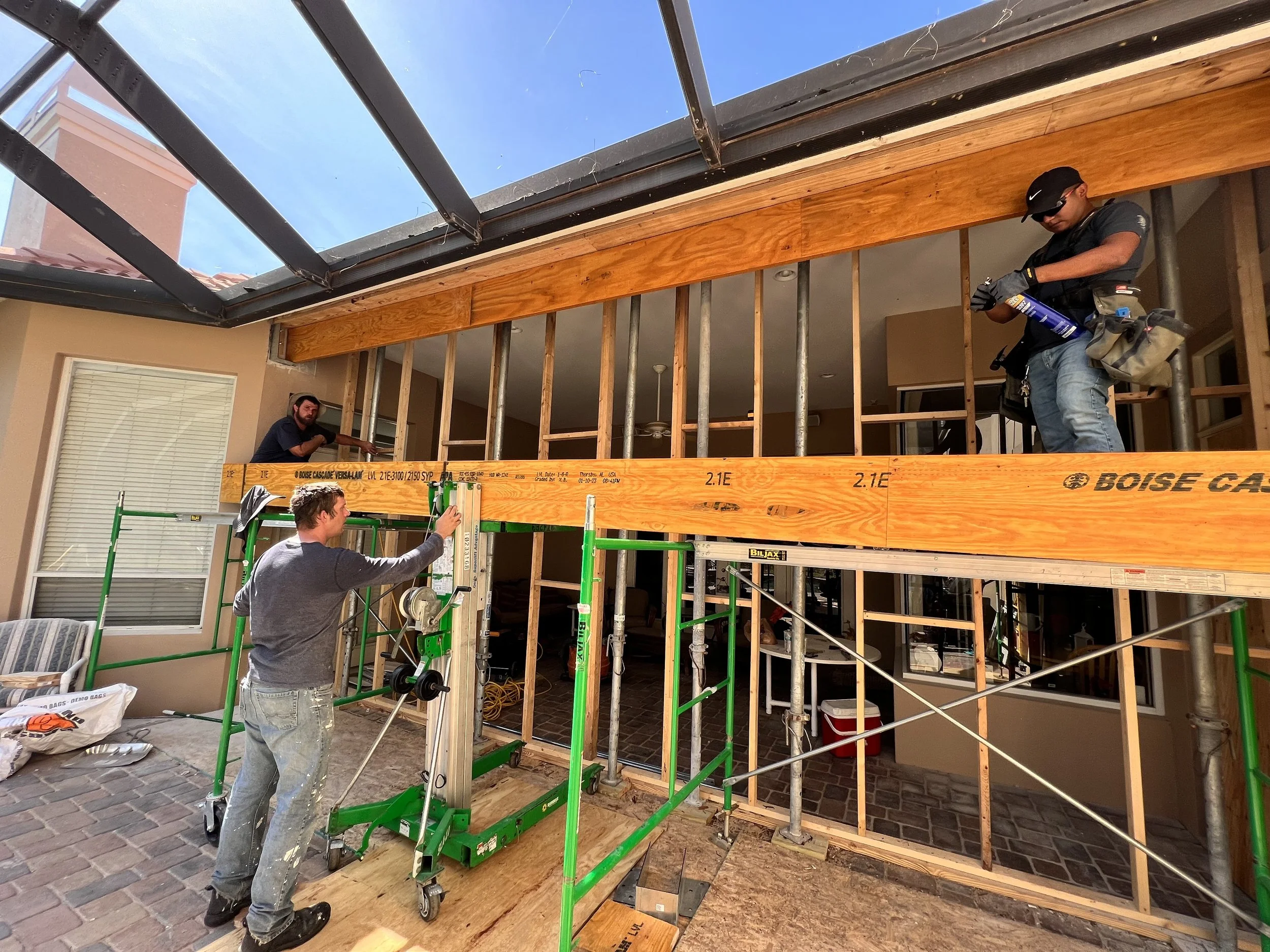 Workers installing a wooden beam on a second-story balcony under construction, with scaffolding and framing in place.