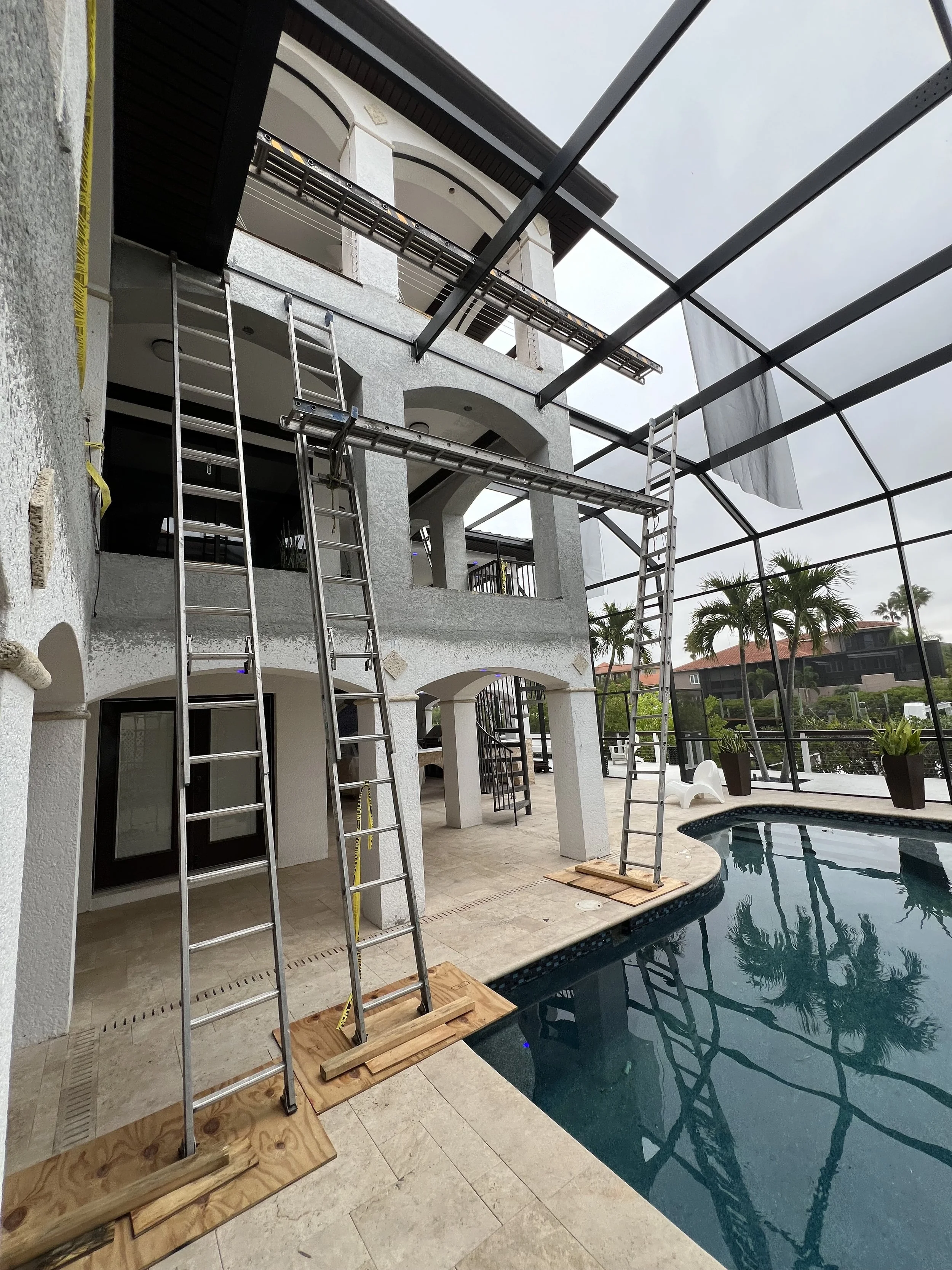 Construction scaffolding and ladders set up around a multi-story house with a swimming pool in the backyard, during daytime with overcast skies.