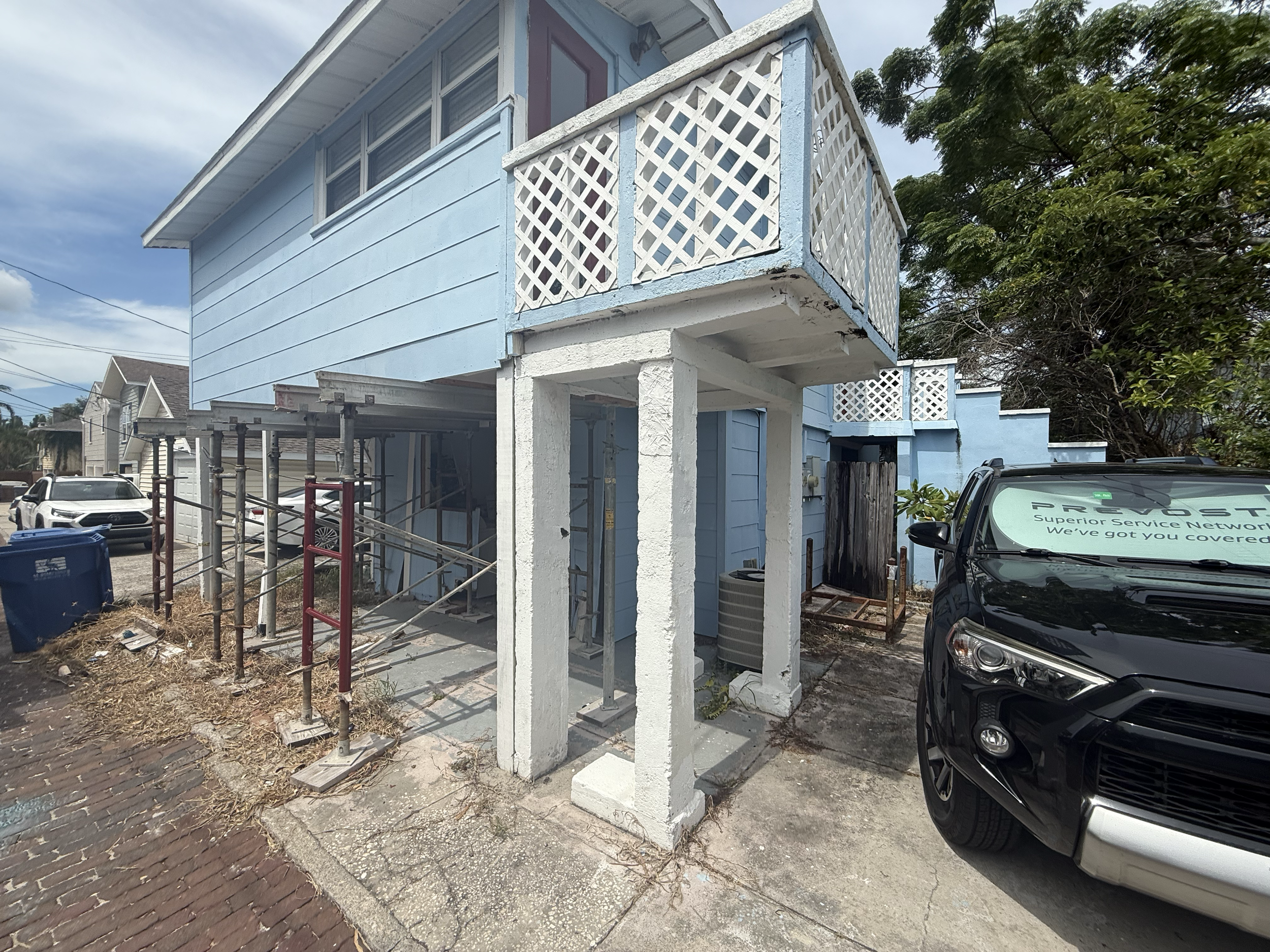 A blue two-story house with a small balcony and lattice railing, parked cars in front, under renovation with scaffolding on the sidewalk, and an air conditioning unit visible outside.