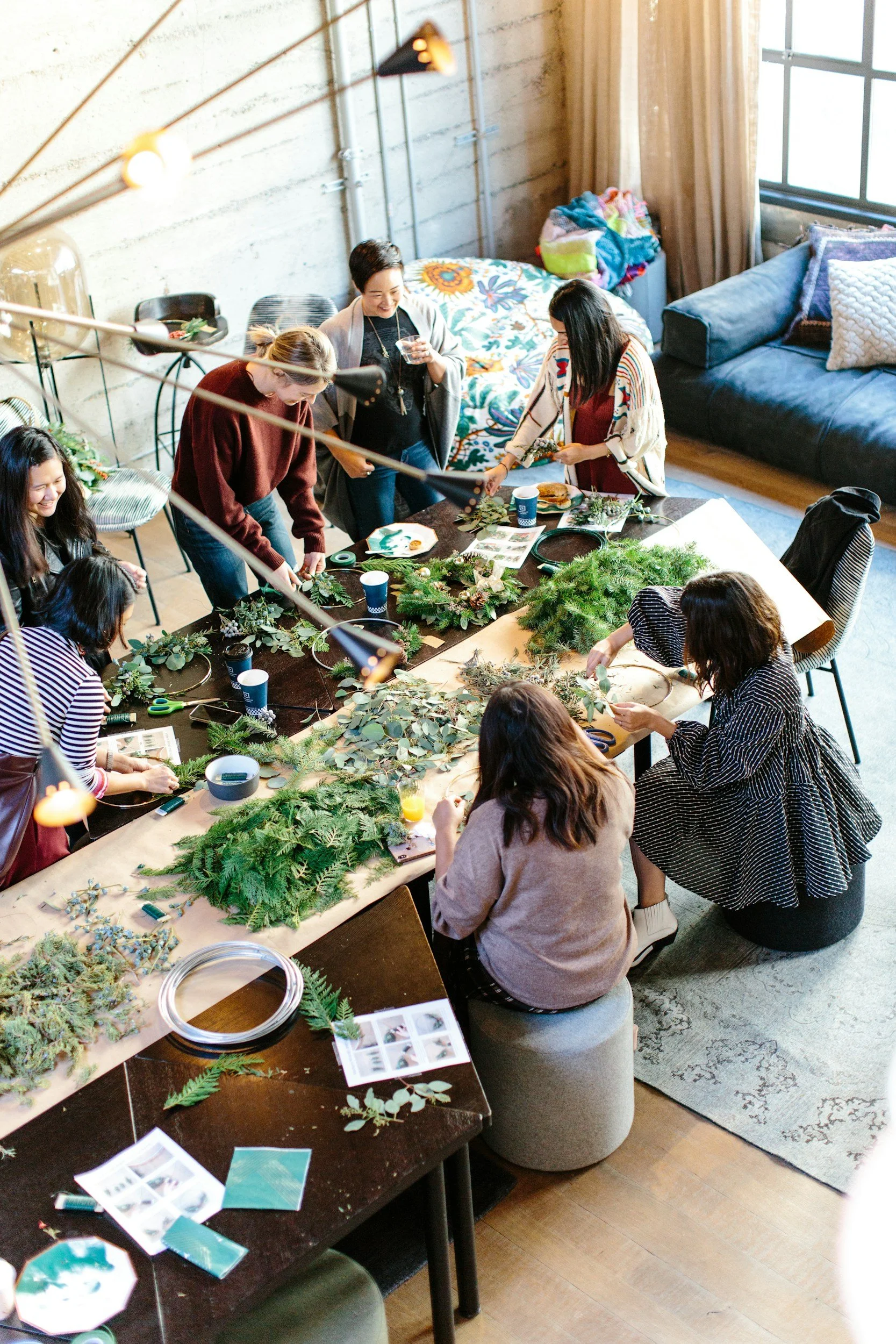 Group of women gathered around a table making holiday wreaths with greenery and decorations in a cozy indoor setting.