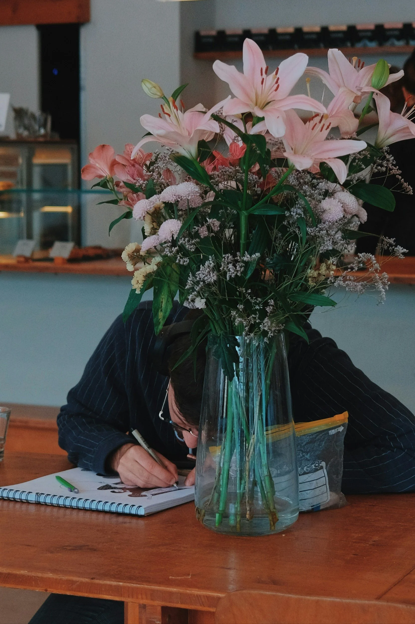 A person writing or drawing at a wooden table, with a large bouquet of pink lilies and other small flowers in a glass vase in front of them.