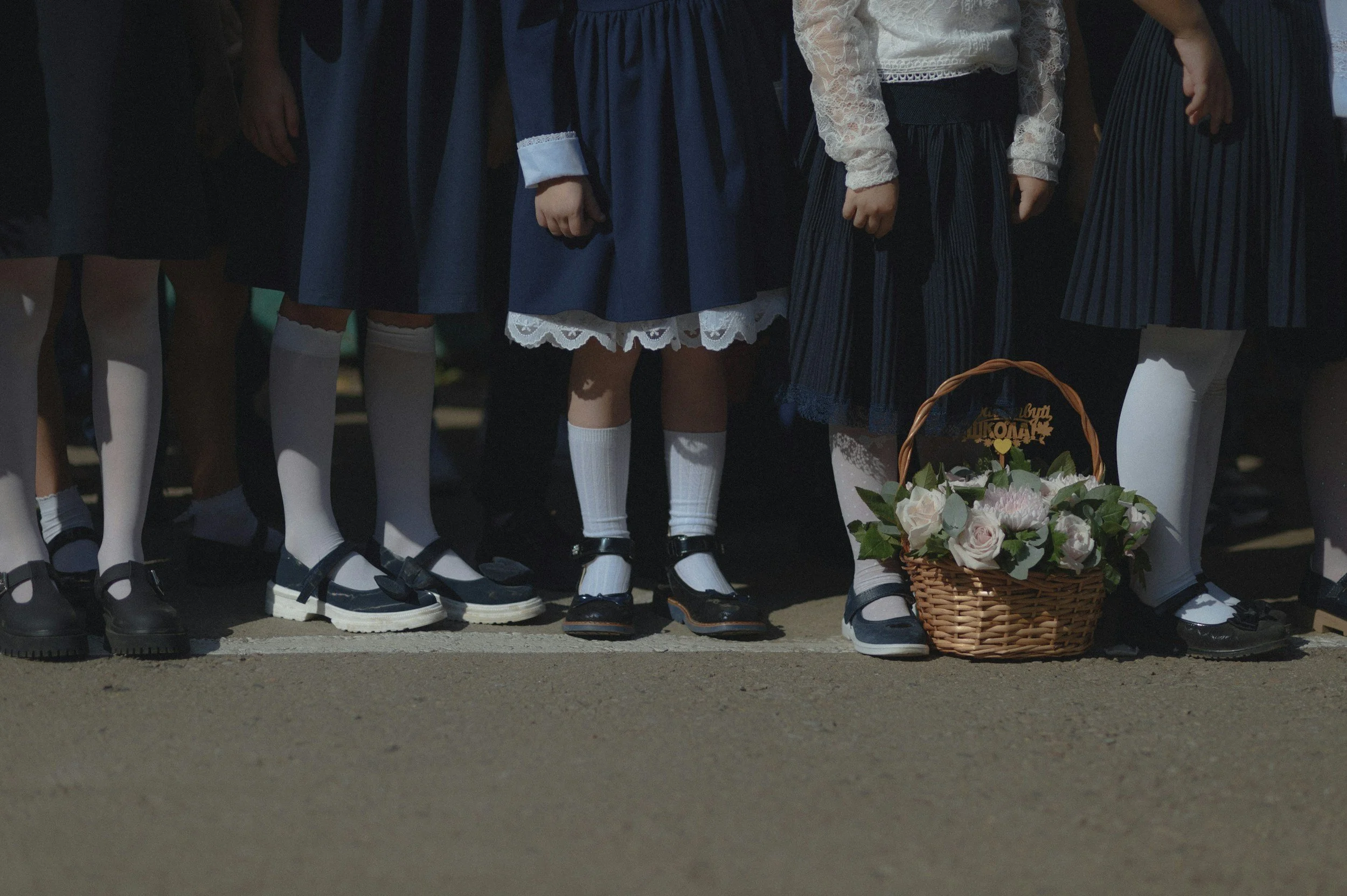 Children dressed in traditional attire standing in a row with white socks and black shoes, and a basket of flowers on the ground.
