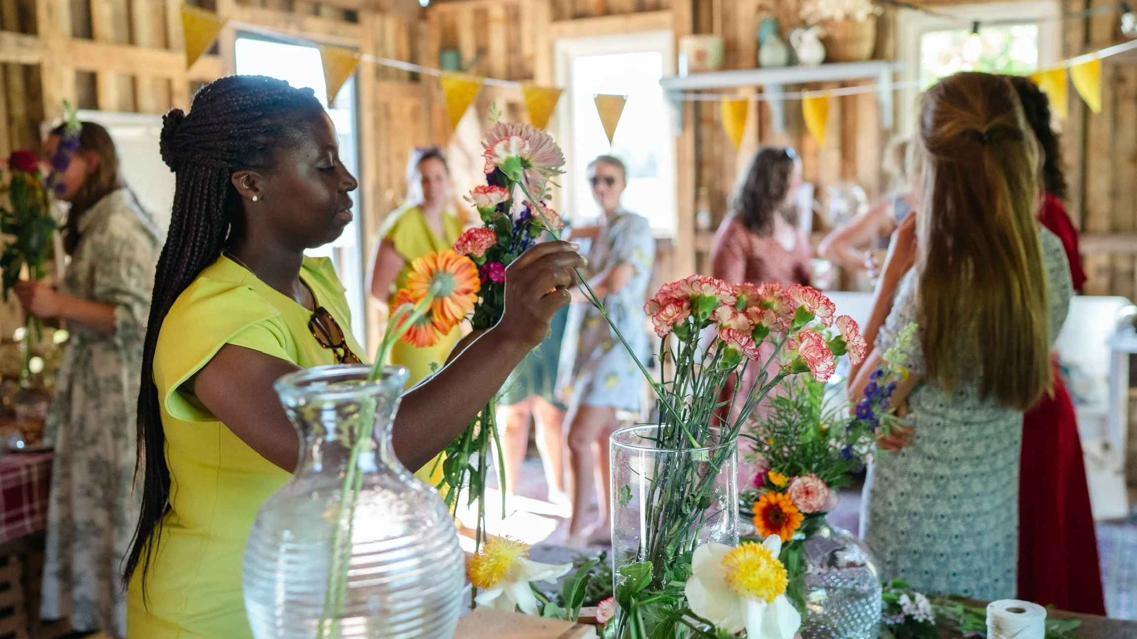Women arranging flowers at a floral display booth during a gathering or event inside a rustic wooden building.