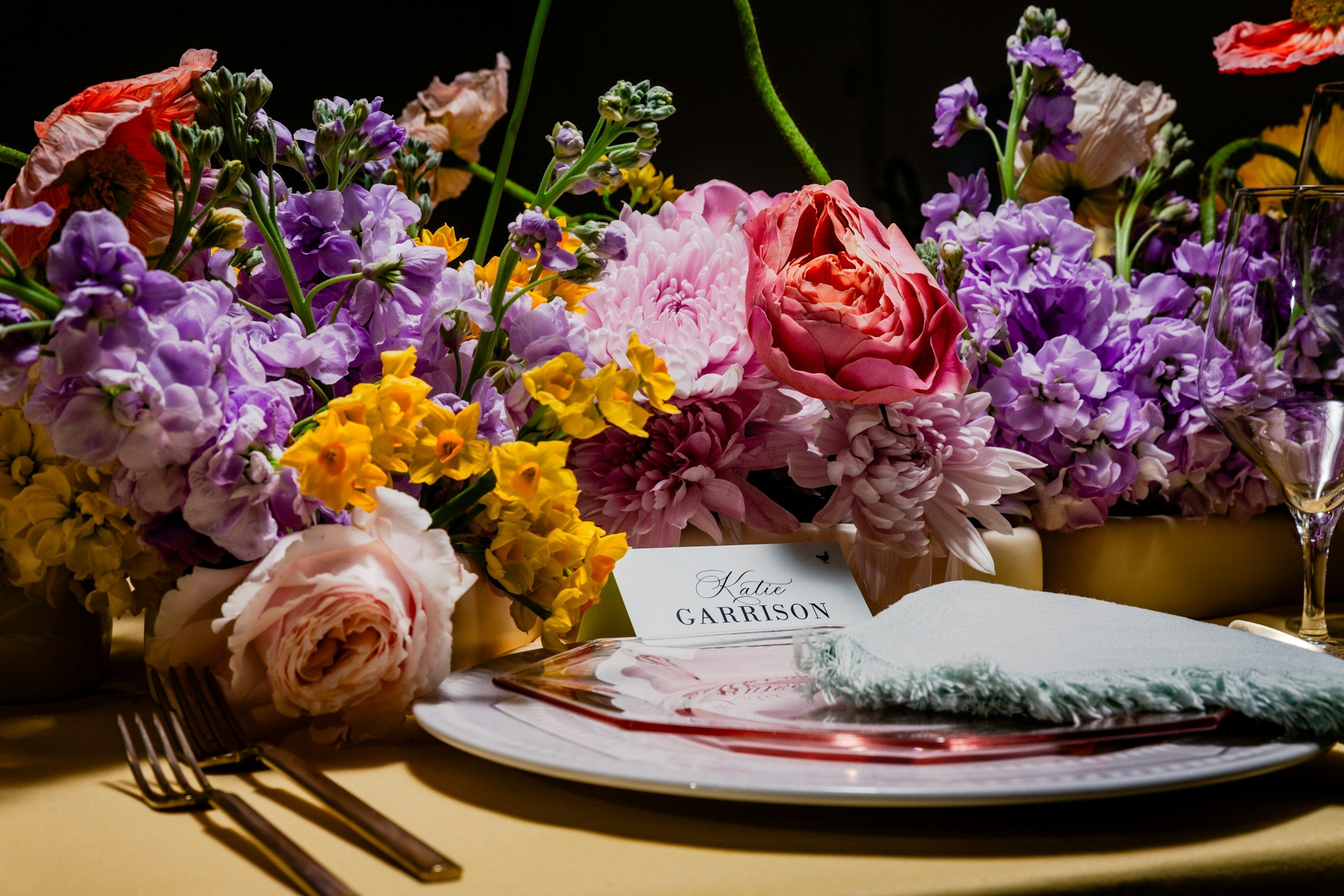 A floral arrangement with various colorful flowers including pink, purple, yellow, and peach blooms, displayed on a table for an event, featuring a name tag for Katie Garrison.