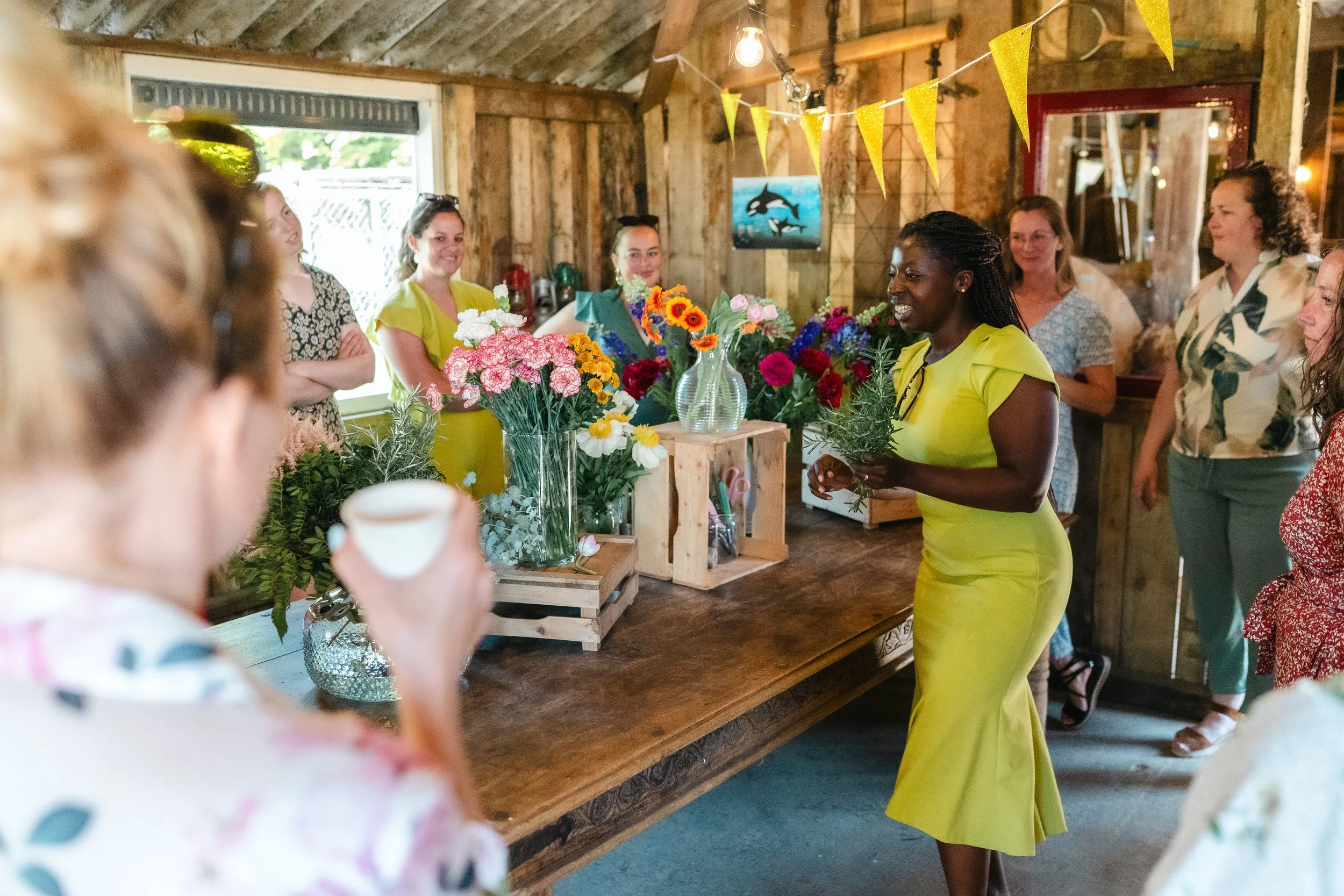 A woman in a yellow dress walking past a table with colorful flower arrangements, inside a rustic wooden room decorated with yellow banners and a picture of dolphins on the wall. Several women are gathered around, some smiling and observing her.