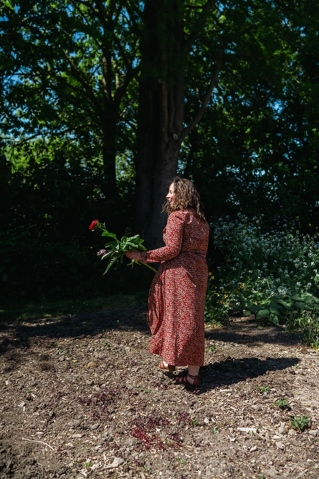 A woman with curly hair wearing a red floral dress holds a bouquet of flowers outdoors on a dirt path with green trees and foliage in the background.