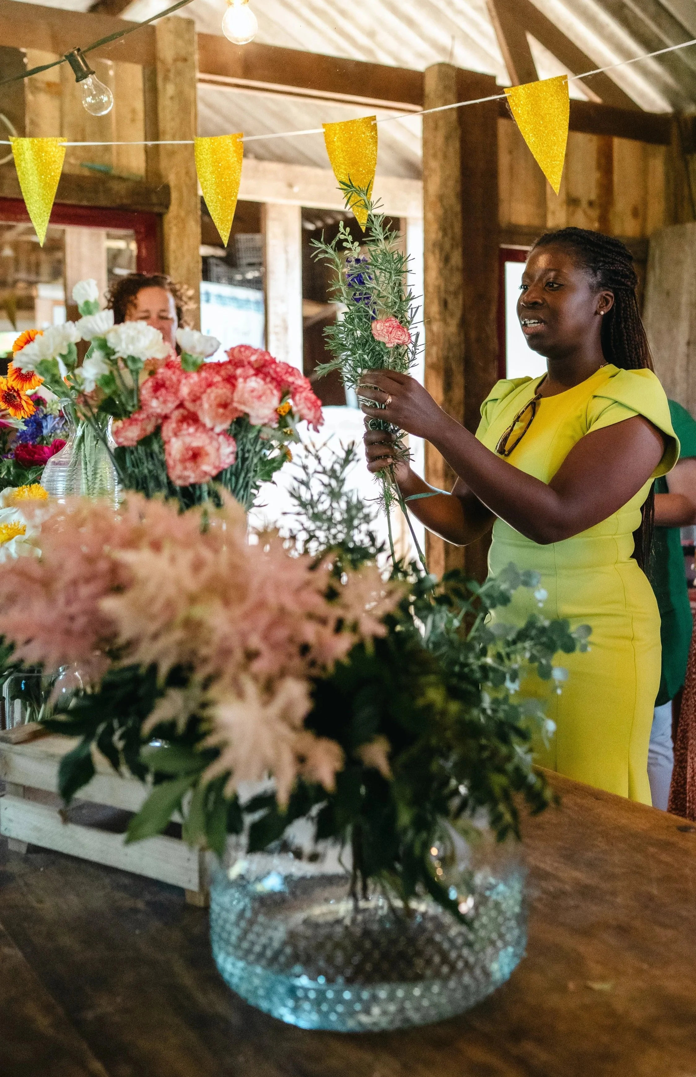 A woman in a yellow dress arranging flowers at a table decorated with various colorful flowers, inside a rustic barn with wooden beams and string lights.
