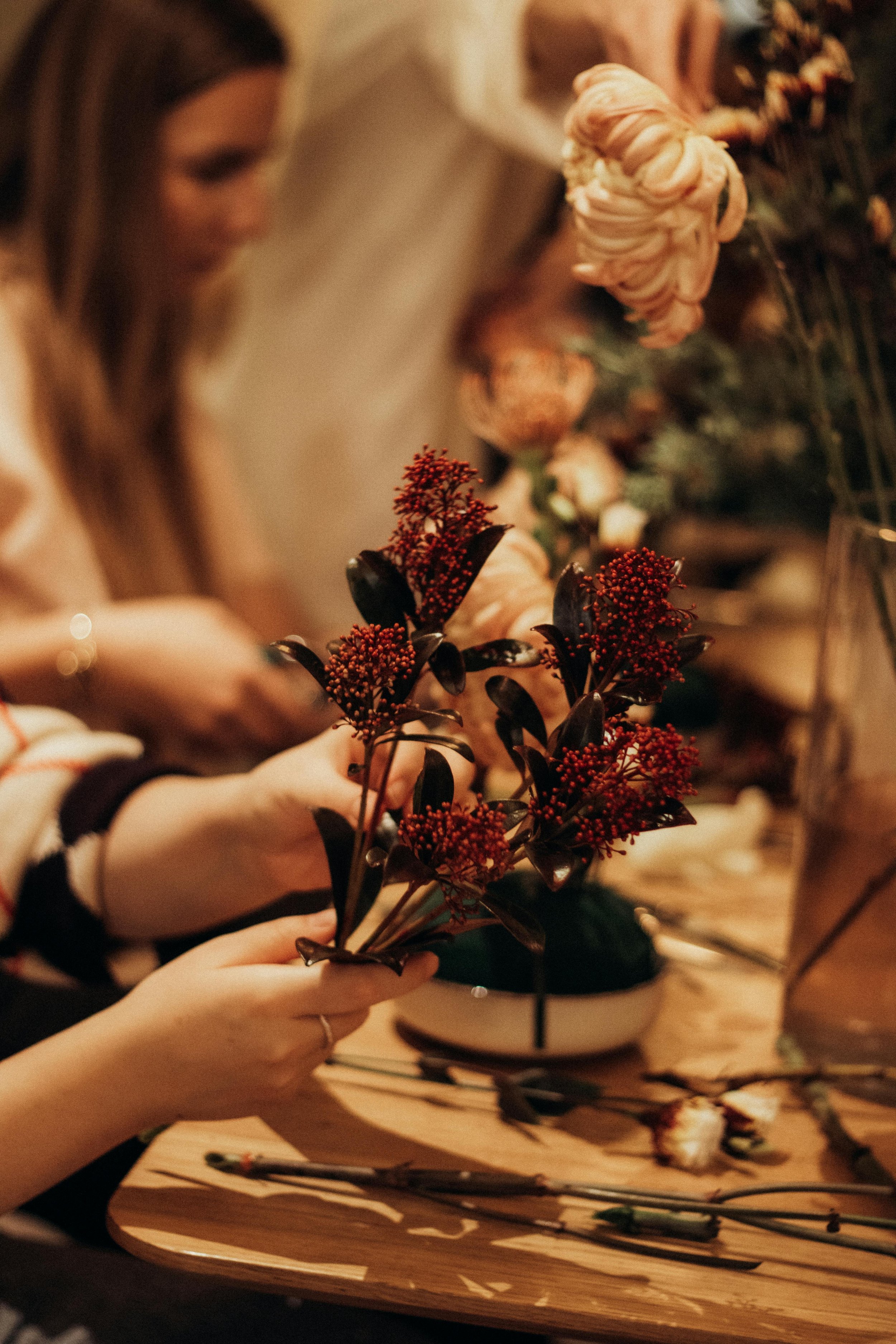 People arranging red and dark-colored flowers on a wooden table, with a blurred woman in the background.