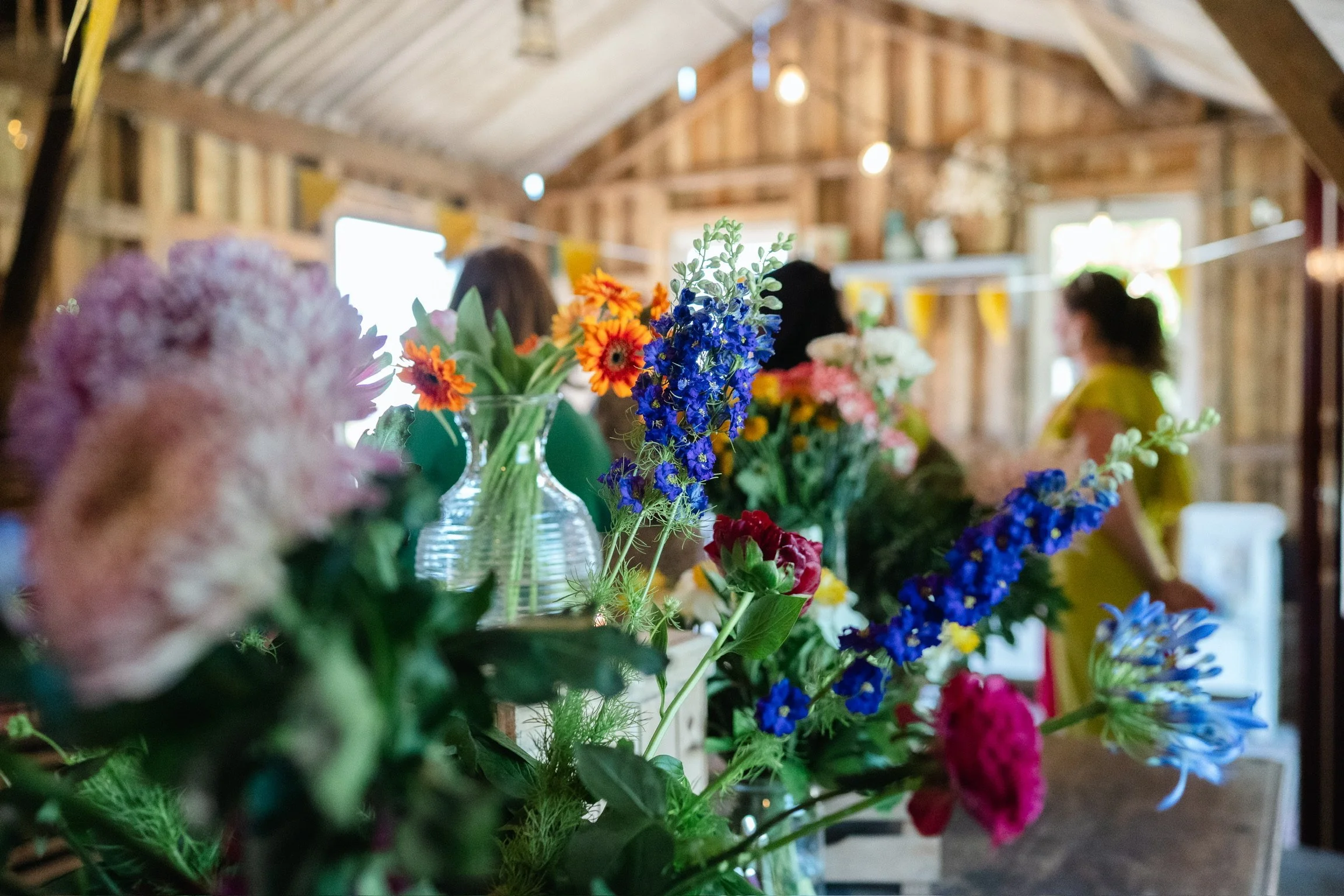 Colorful flowers displayed on a table inside a rustic barn with wooden walls and ceiling, with people in the background.