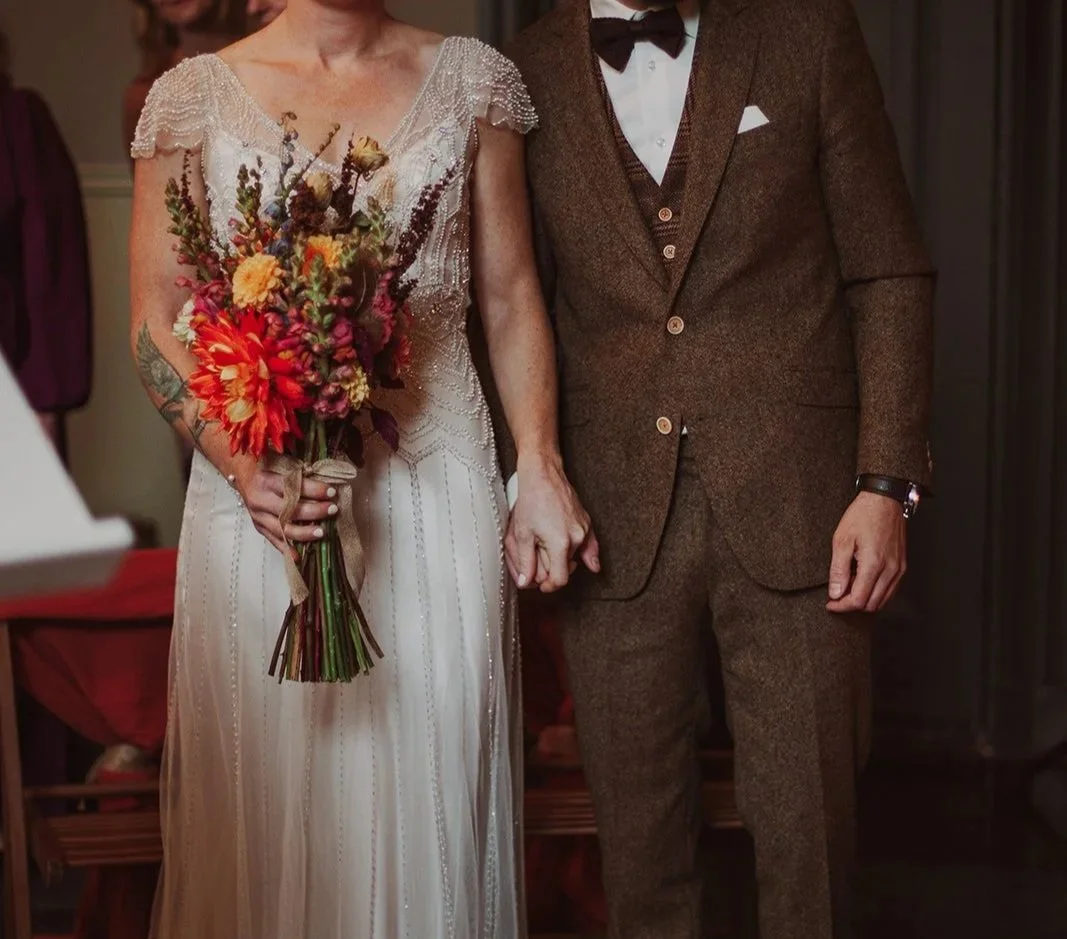 A bride and groom holding hands during their wedding ceremony, with only their torsos visible. The bride is in a vintage-style white wedding dress holding a colorful bouquet, and the groom is in a brown suit with a white shirt and black bow tie.