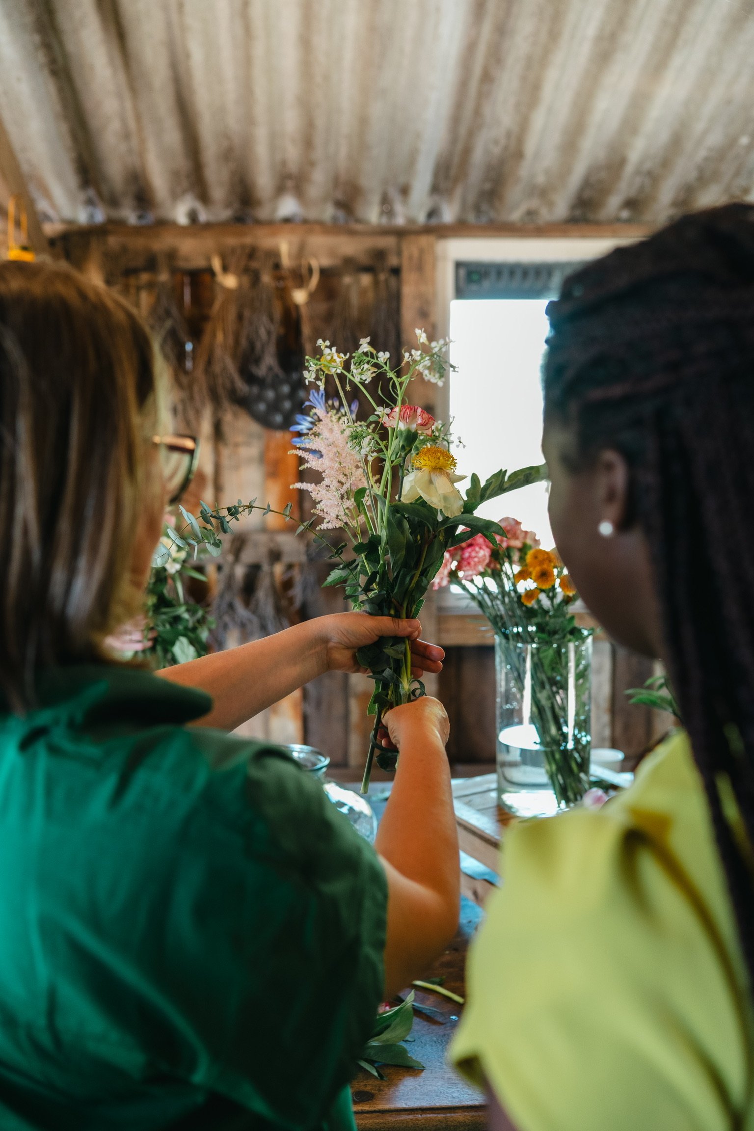 Two women are exchanging a bouquet of colorful flowers inside a rustic wooden room with a tin roof and a window in the background.