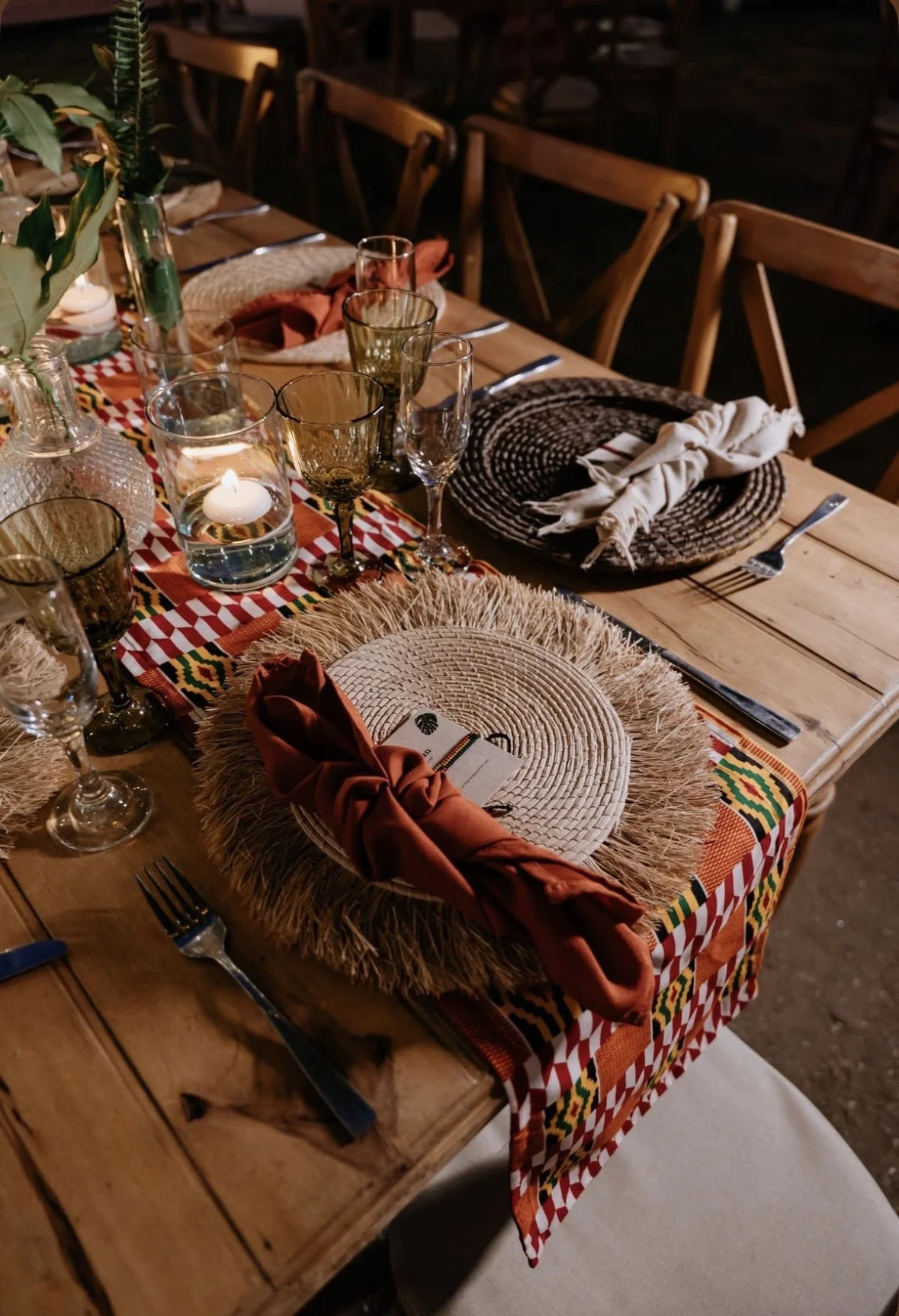 A rustic dining table set for a dinner with woven placemats, cloth napkins, wine glasses, candles, and a colorful patterned table runner.