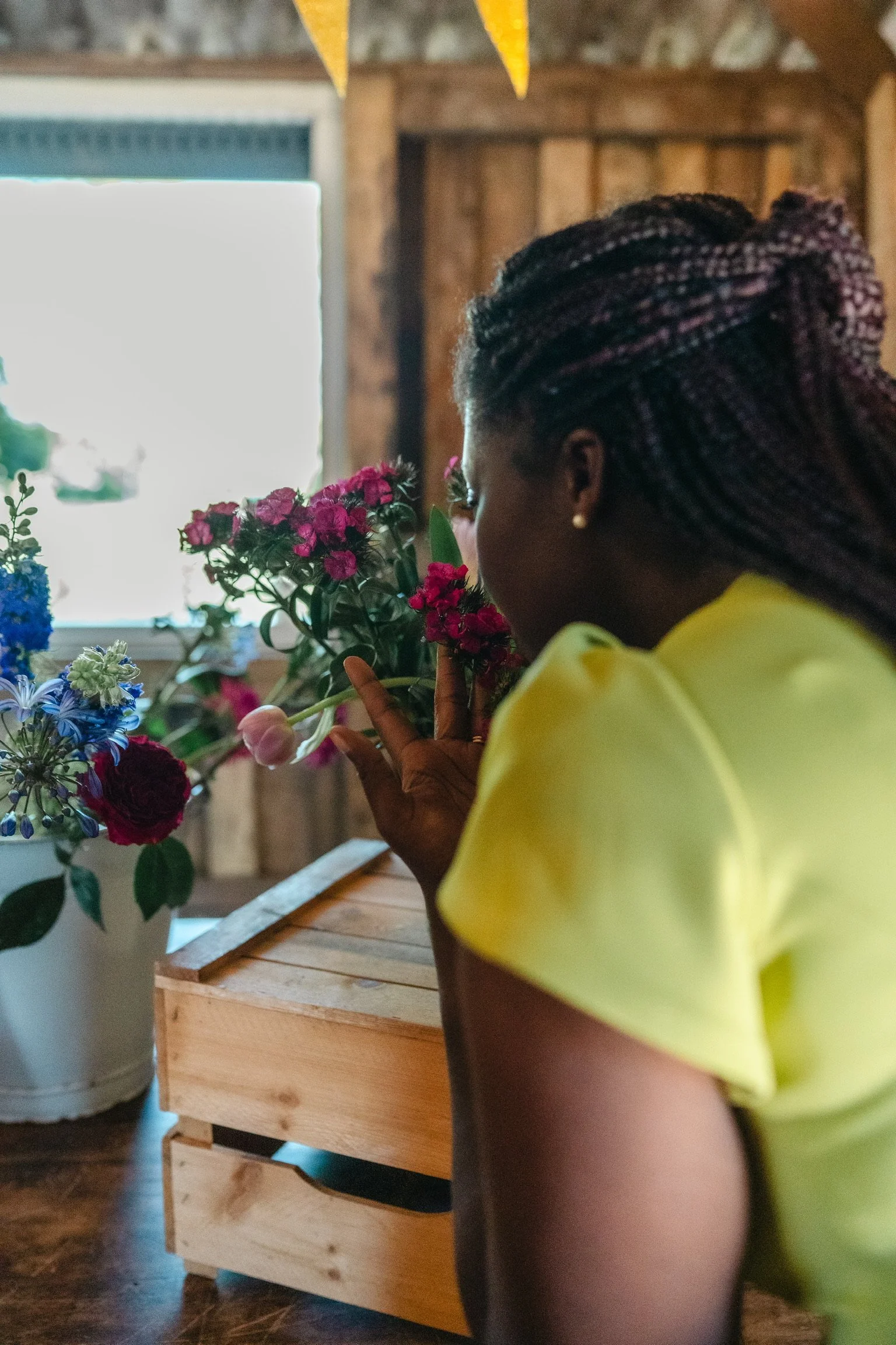 A woman with braided hair and earrings smells a pink flower at a flower shop, with a window and wooden interior in the background.