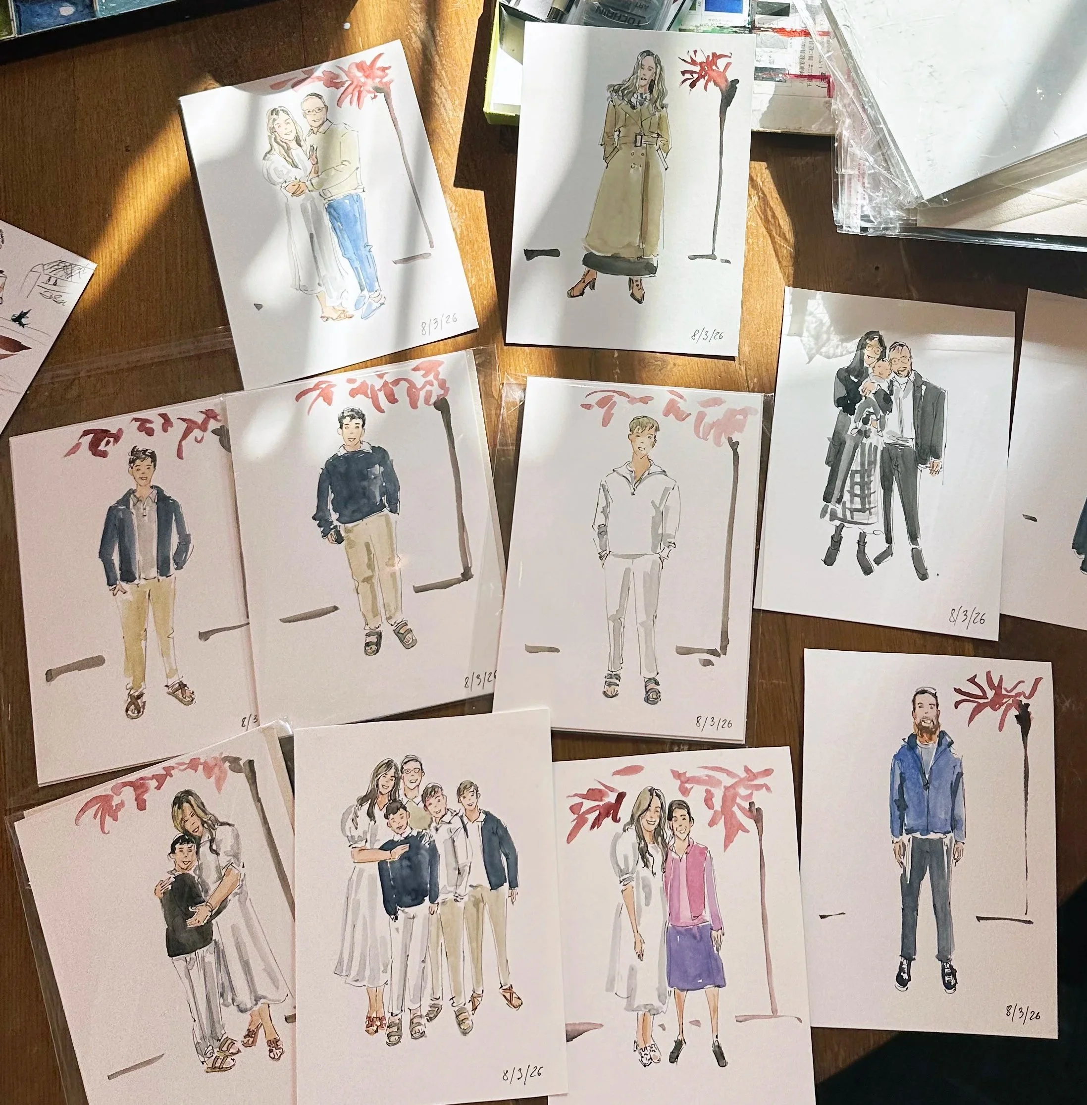 Collection of watercolor fashion sketches of people, some in pairs, on a wooden table with sunlight and stationery around.