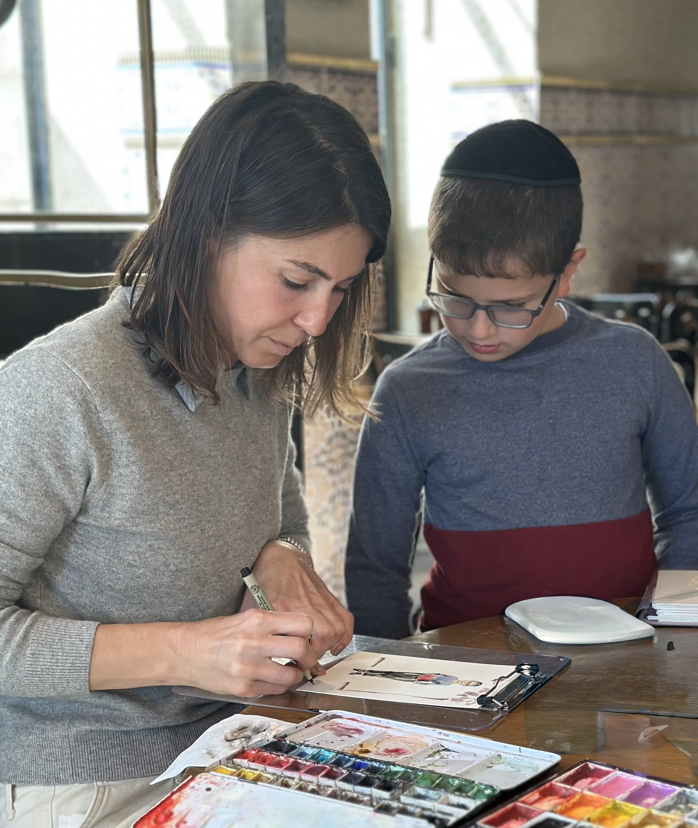 A woman and a boy working together on a watercolor painting at a table in a cozy indoor setting.