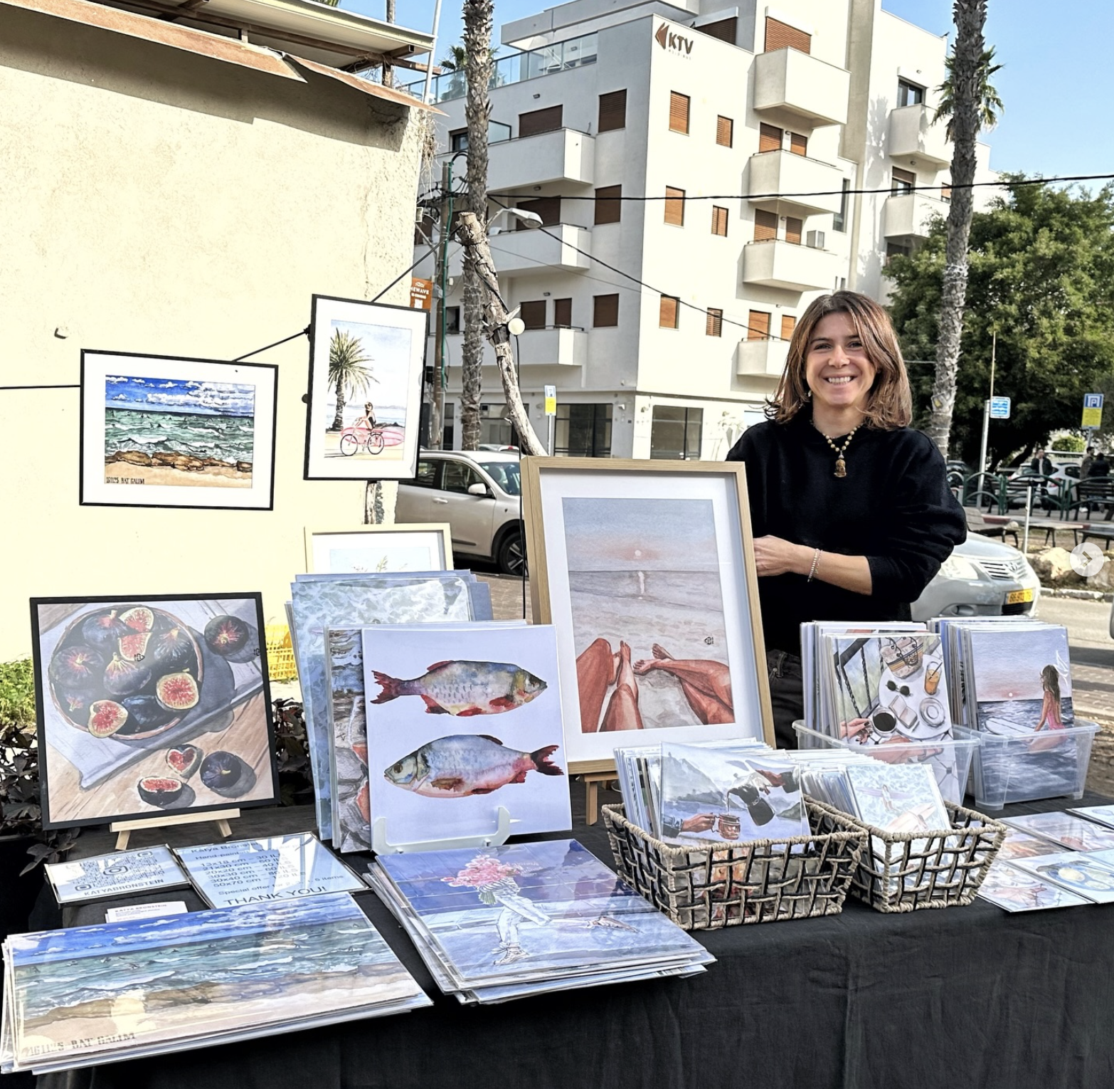 A woman at an outdoor art market displaying her various framed artworks, including paintings of the beach, fish, and food, on a table with baskets of postcards.