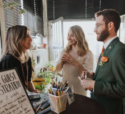 An live event artist and a newlywed couple indoors, engaged in conversation. All three people are smiling. The scene is bright and welcoming.