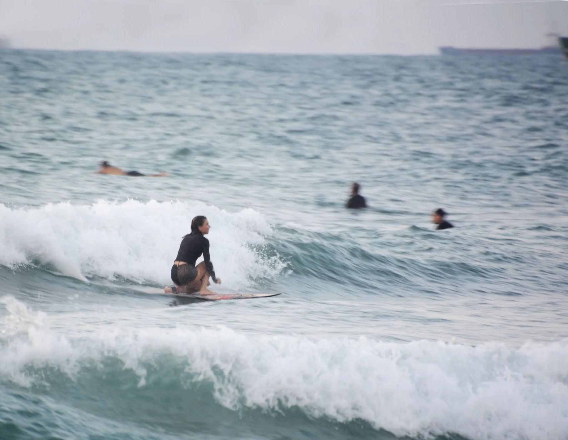 A woman surfing on a small wave with several people swimming and floating in the background in the sea.