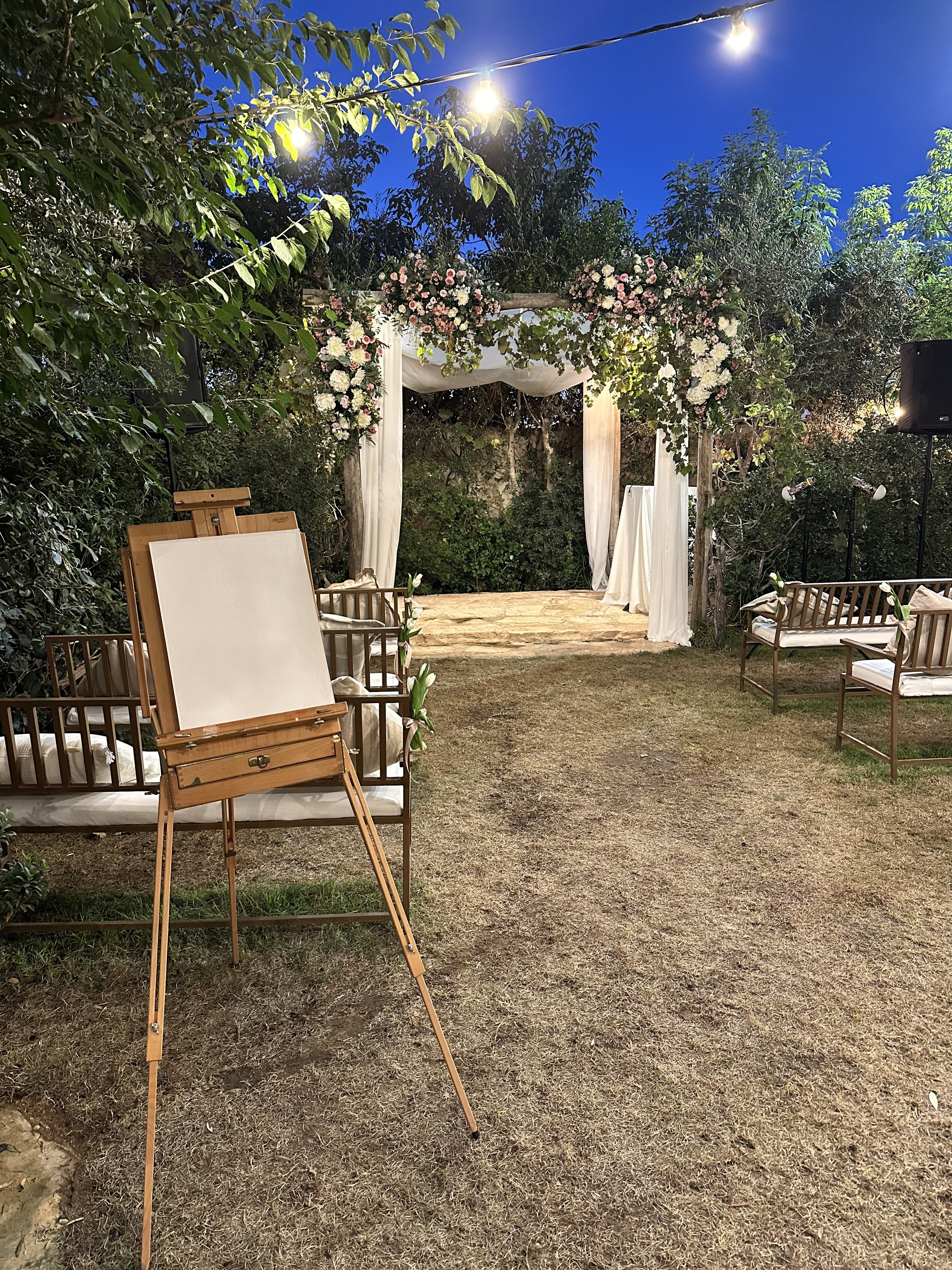 Outdoor wedding ceremony setup in a garden with a wooden arch decorated with pink and white flowers, surrounded by chairs with white cushions, under string lights at dusk.