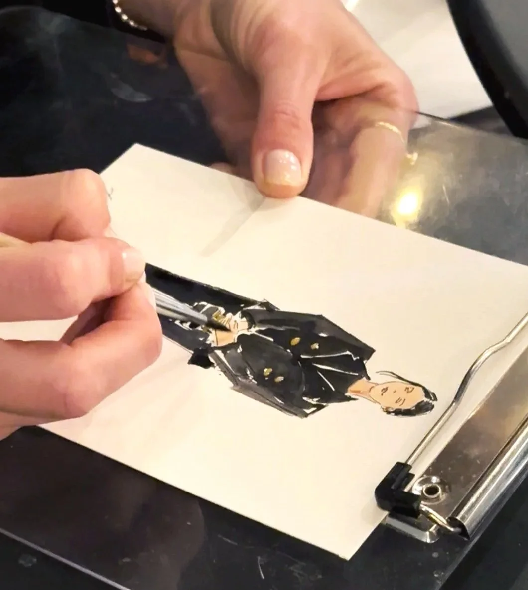 Person uses a brush to paint a fashion illustration of a woman in a black dress on paper.
