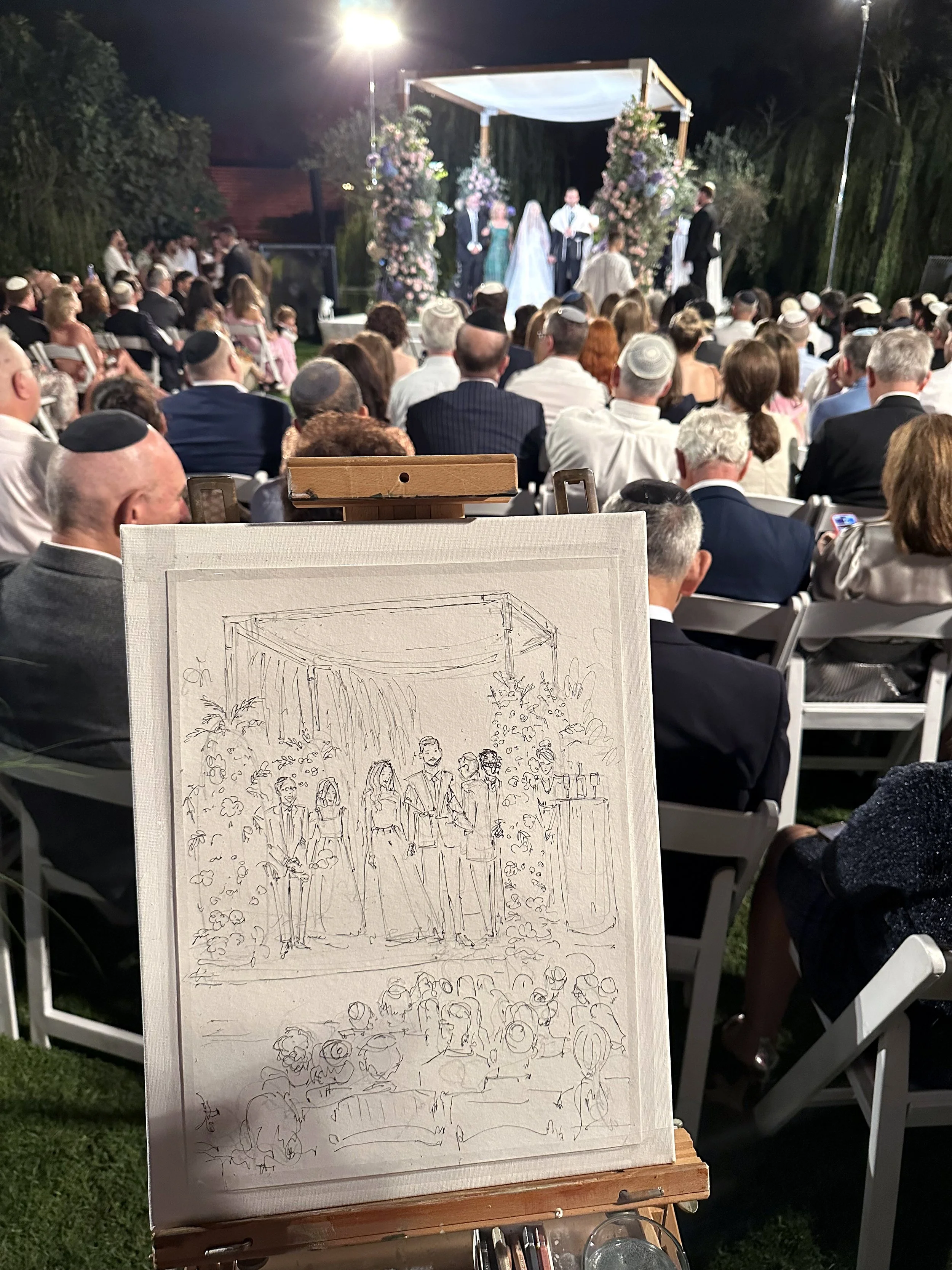 A sketch of a wedding ceremony on an easel in front of an outdoor wedding with guests seated and a couple walking down the aisle, with a decorated arbor and floral arrangements, at night.