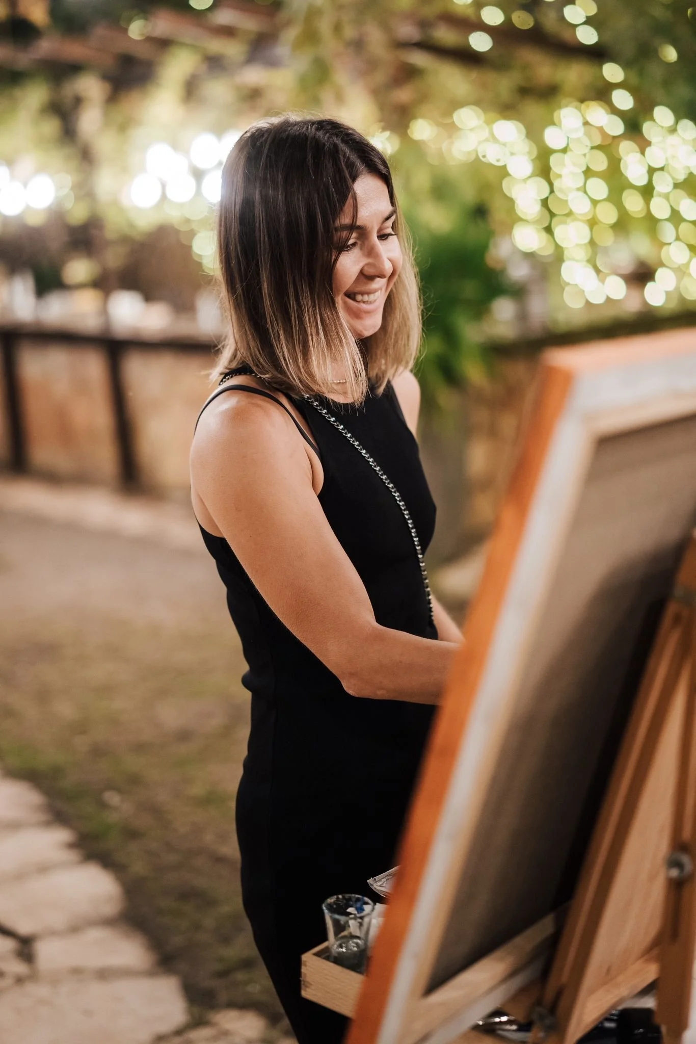A woman in a black dress painting outdoors at night, smiling while working on a canvas set on an easel.