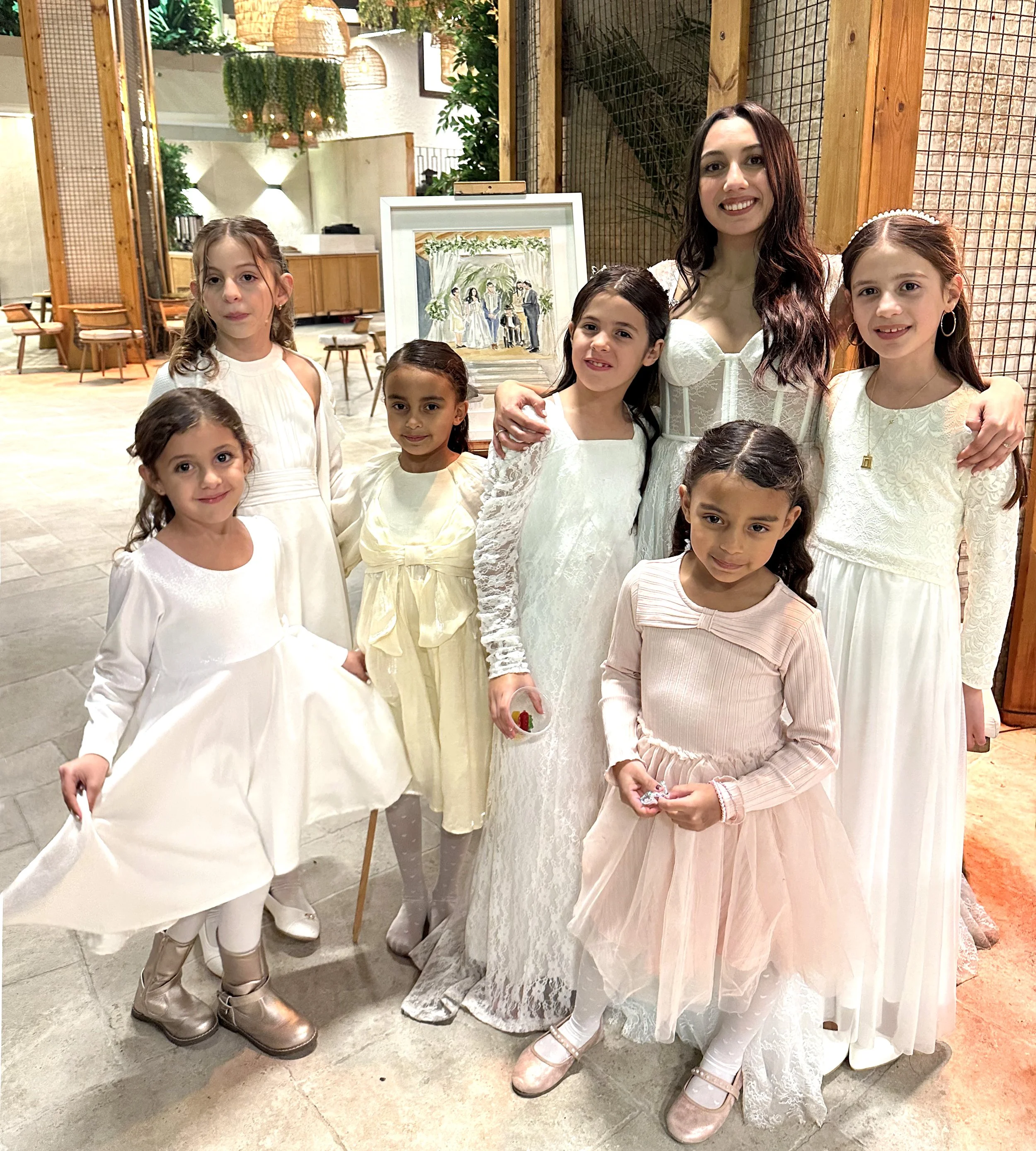 Group of young girls and woman dressed in white and pastel dresses, posing indoors at a celebration or special event.