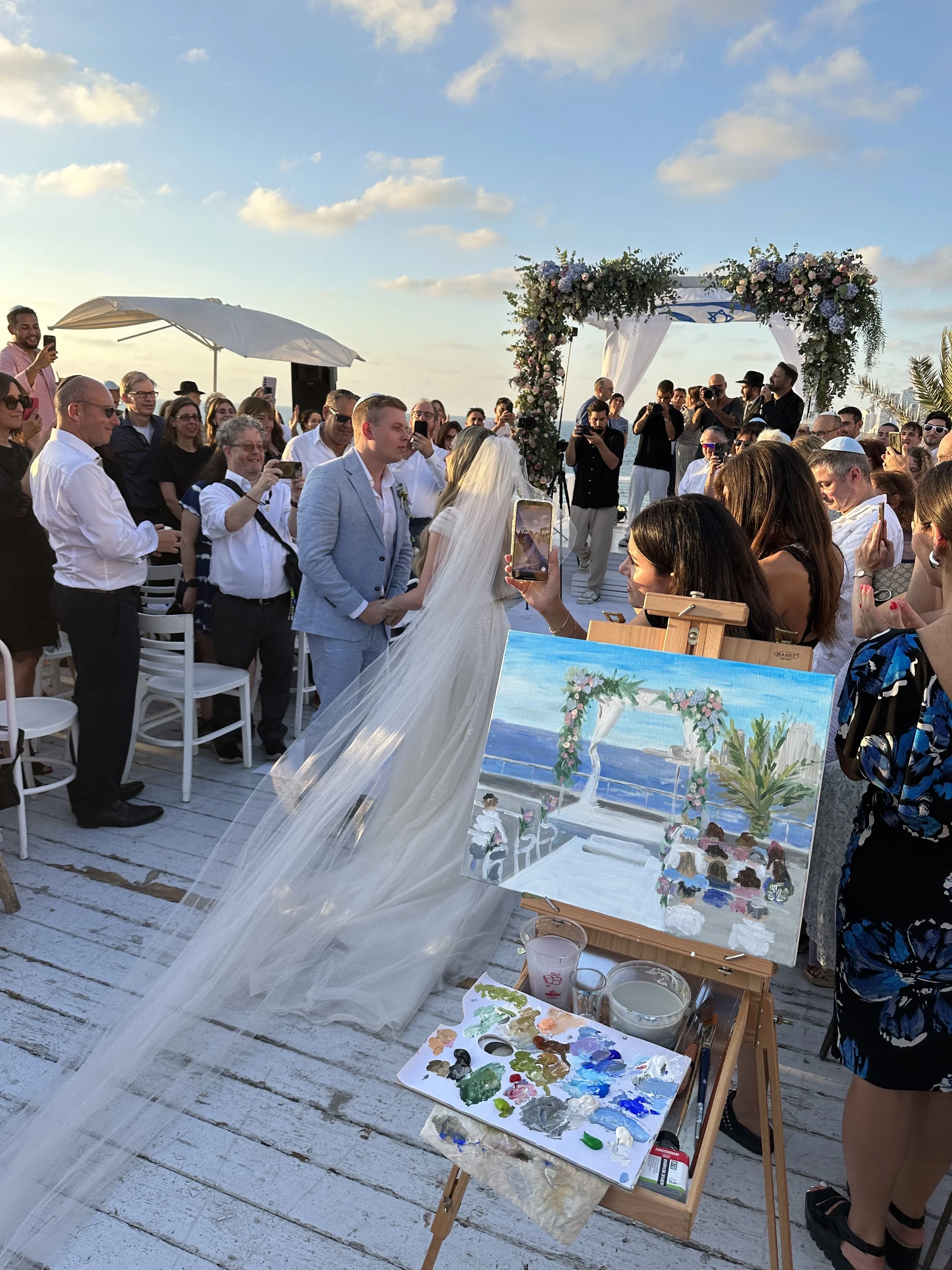 A wedding ceremony on a wooden deck with a bride and groom facing each other under a floral arch, surrounded by guests taking photos. An artist's painting of the scene is displayed on an easel in the foreground, along with a palette and painting supplies.