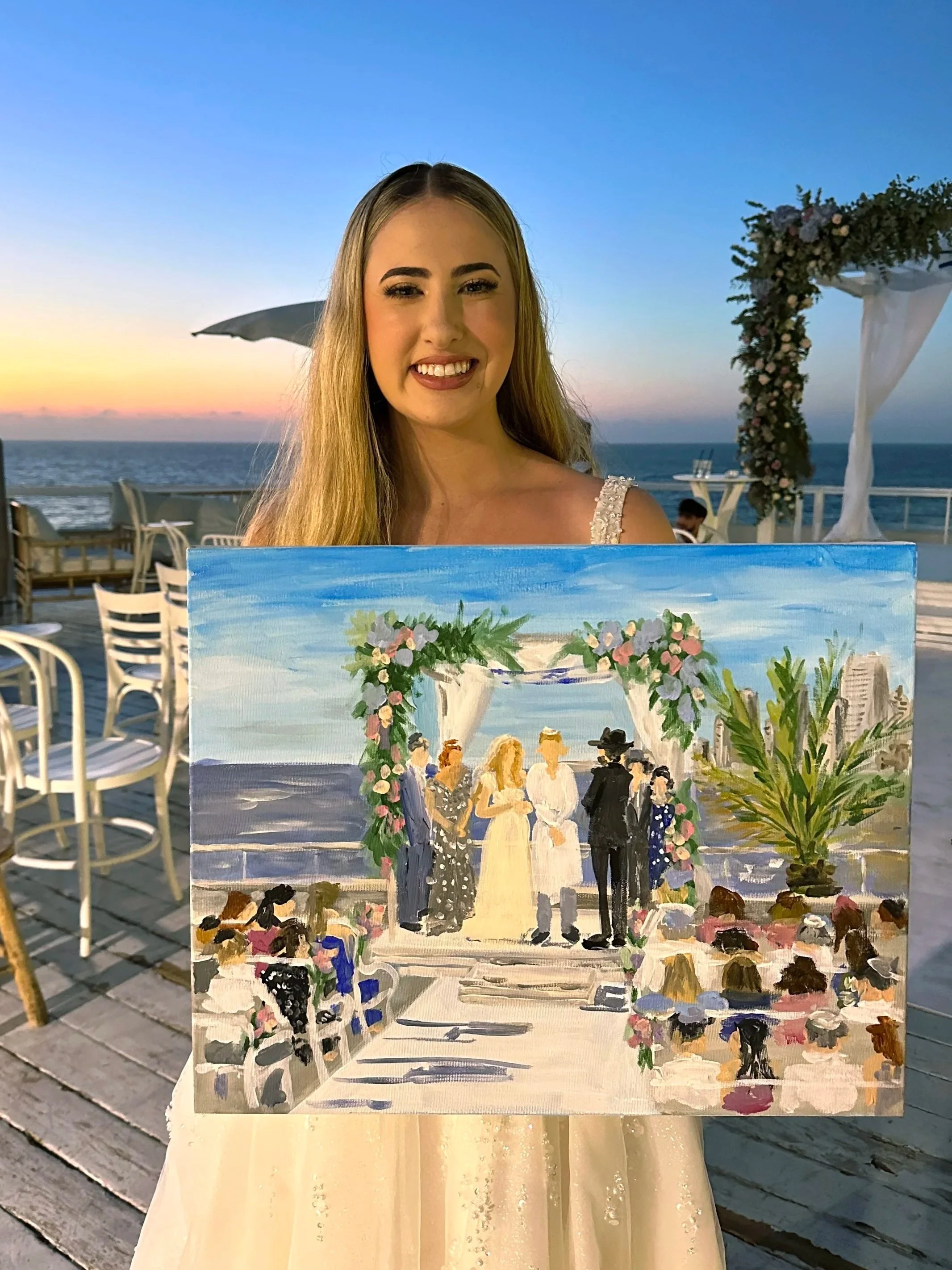 A young woman with long blonde hair holding a painting of a wedding ceremony on a beach during sunset, with chairs and a floral arch in the background.