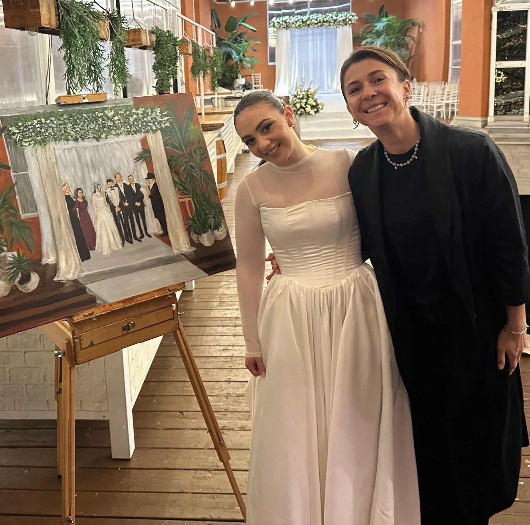 A young bride in a wedding dress standing next to an older woman artist in black, smiling at a wedding reception with a painting of a wedding scene on an easel beside them.