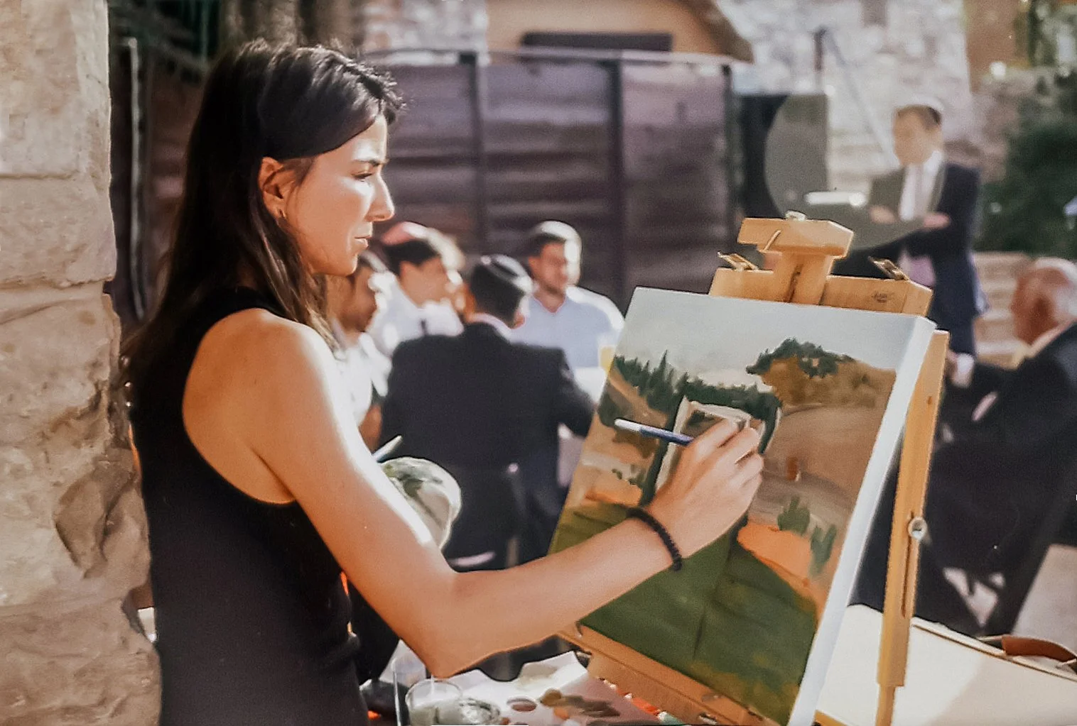 A woman painting a landscape on a canvas at an outdoor event, with people gathered in the background.