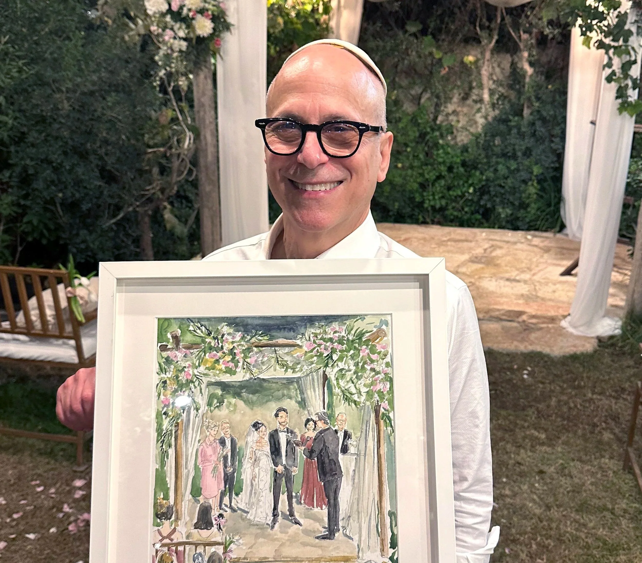 A man wearing glasses and a white shirt is smiling while holding a framed watercolor painting of a wedding scene, featuring a couple exchanging vows under a floral arch surrounded by guests at an outdoor wedding.