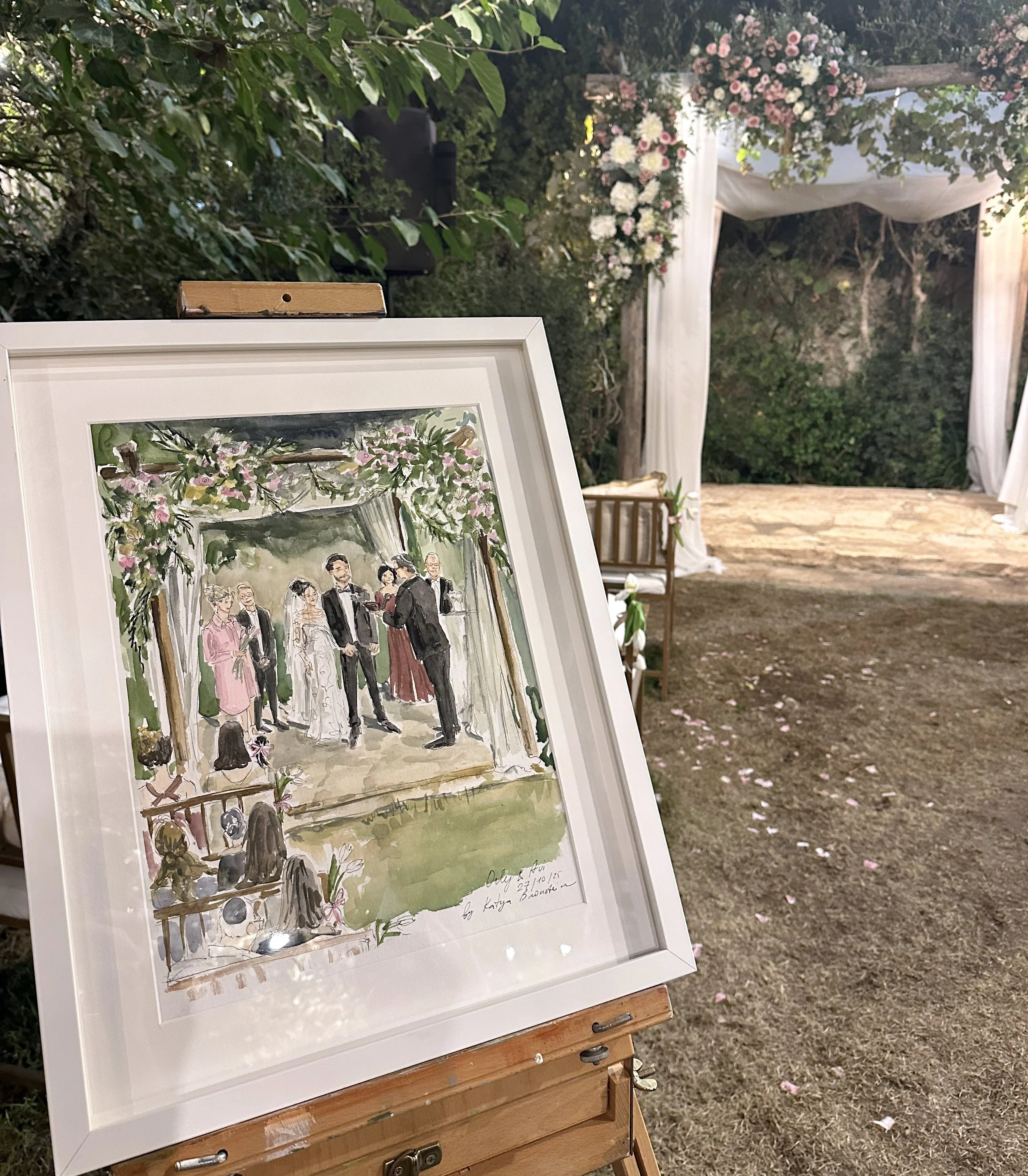 A framed watercolor painting of a wedding ceremony is displayed on an easel outdoors. The scene in the painting shows a bride and groom standing under a floral arch with bridesmaids, groomsmen, and guests gathered around. The real location in the background features a decorated outdoor altar with floral arrangements and a canopy, surrounded by trees and greenery.